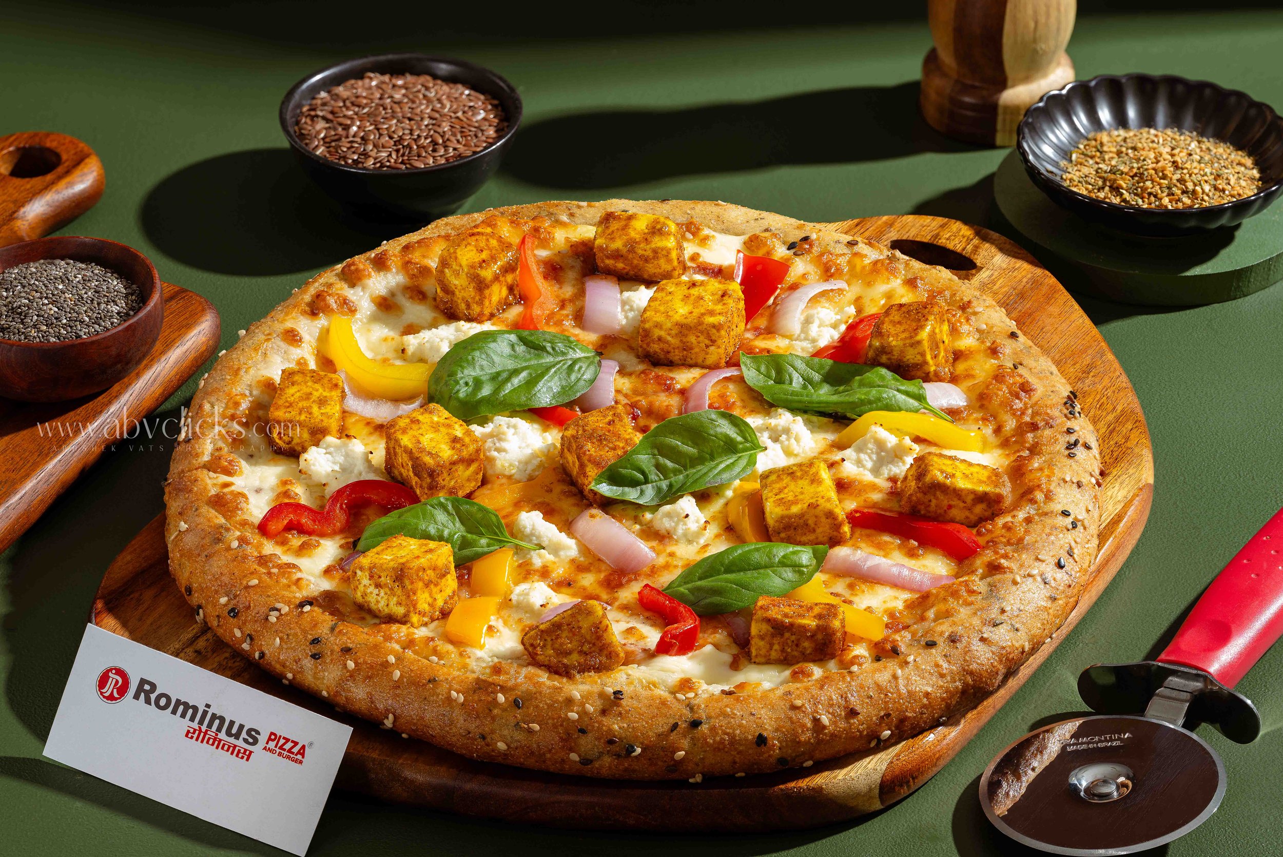 Close-up of a pepperoni pizza with visible toppings of cheese, red, yellow peppers, onions, basil, and tofu cubes on a wooden board, surrounded by small bowls of seeds and spices on a green table.