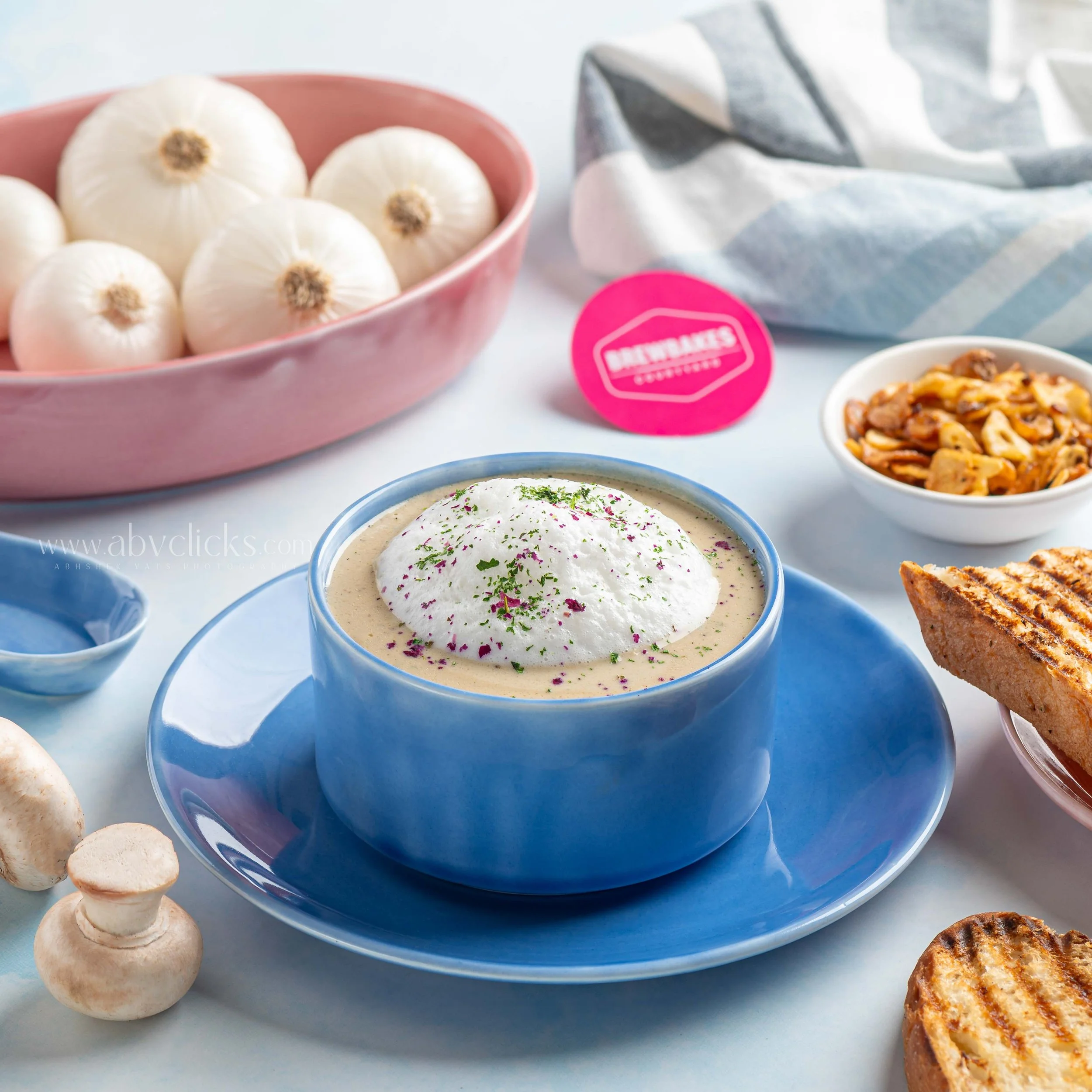 A bowl of white onions, some mushrooms, and a bowl of soup garnished with herbs on a light-colored surface.
