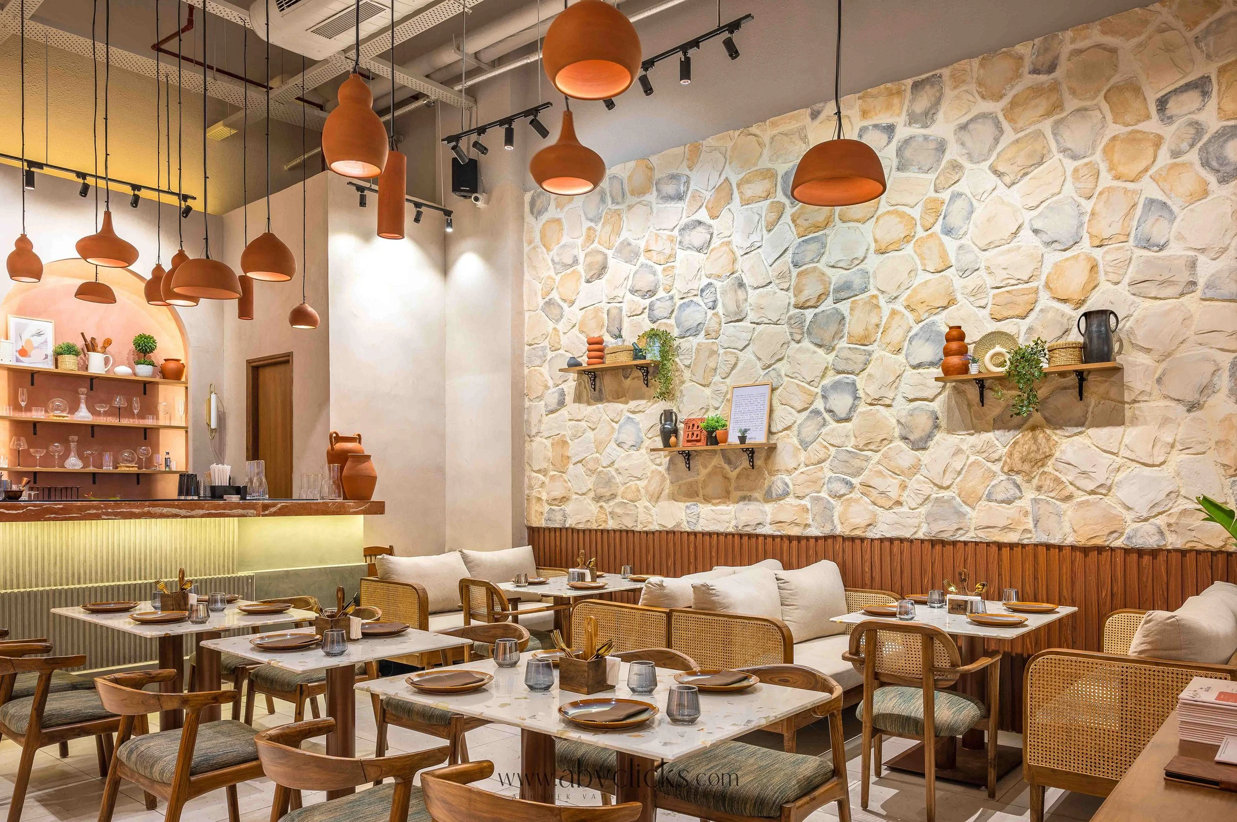 A cozy restaurant interior with a stone accent wall, wooden shelves displaying decorative items, and a ceiling with multiple hanging terracotta-colored pendant lights. The dining area has white marble tables set with plates, glasses, and utensils, su