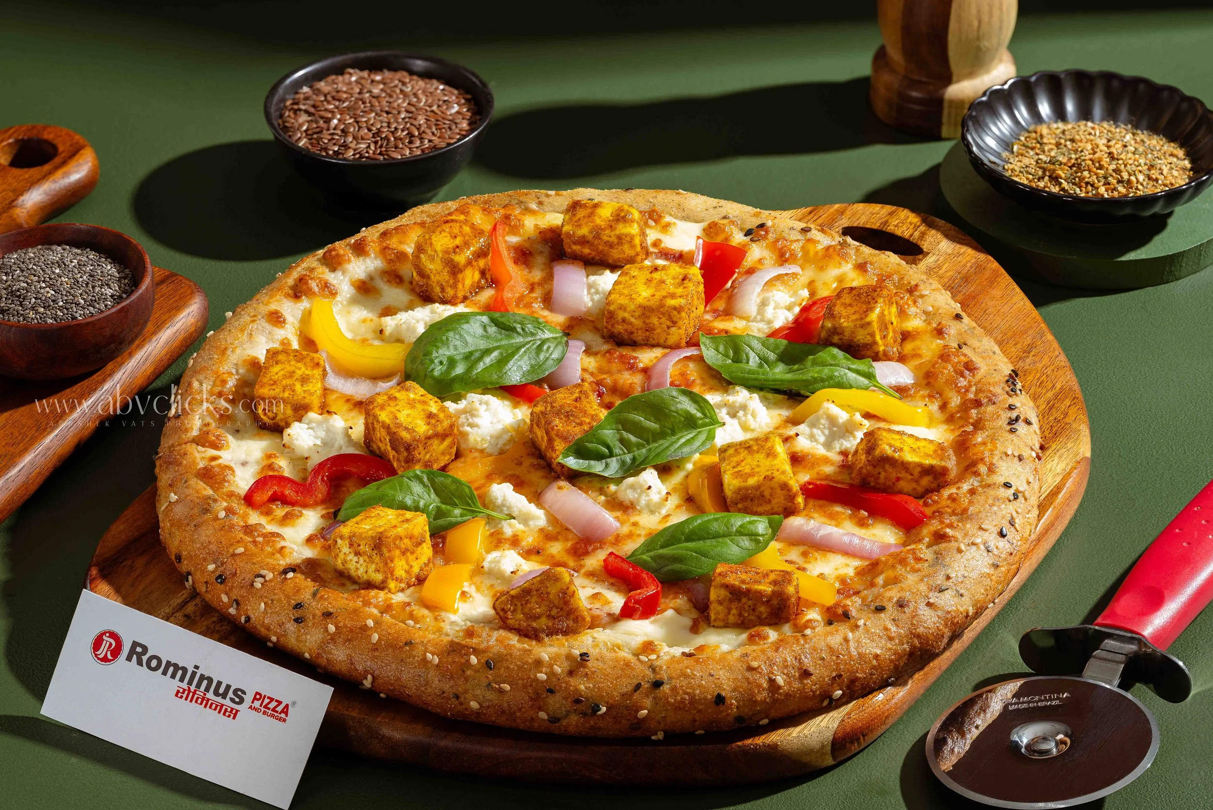 A pizza with tofu, cheese, red, yellow, and white onions, basil leaves, and red peppers on a wooden serving board, surrounded by bowls of flax seeds, sesame seeds, and other spices on a green table.