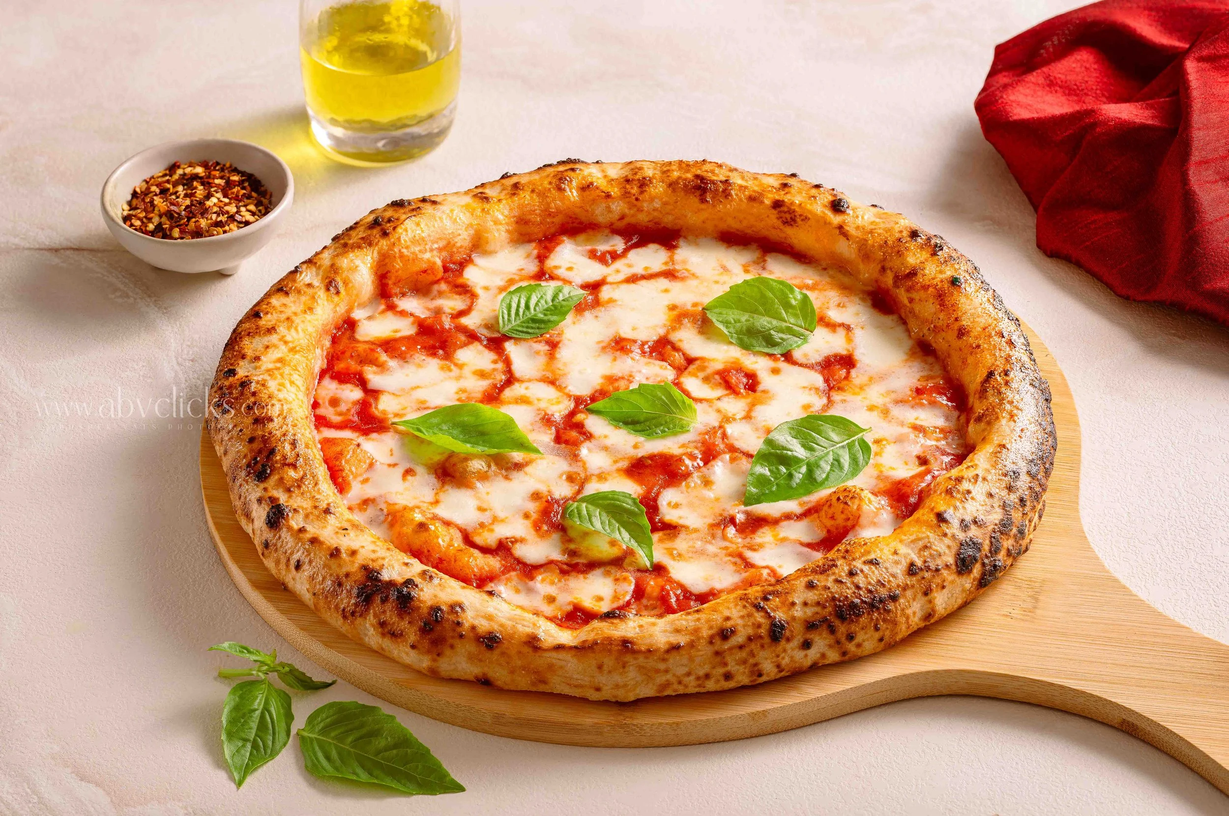 A cheese pizza topped with fresh basil leaves on a wooden pizza peel, with a bowl of crushed red pepper flakes, a glass of yellow beverage, and a red napkin nearby.