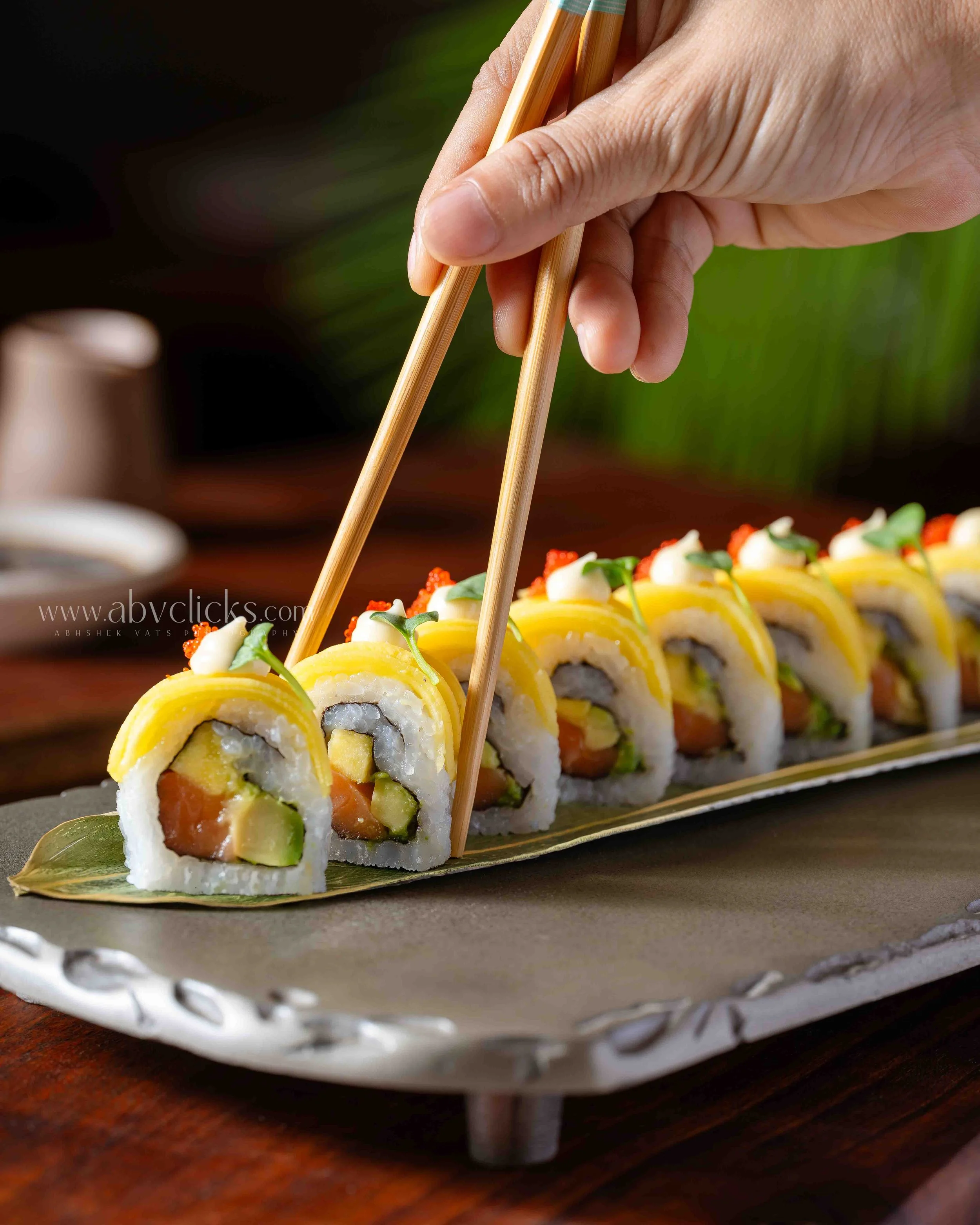 Close-up of a hand using chopsticks to pick up sushi rolls arranged on a decorative platter.