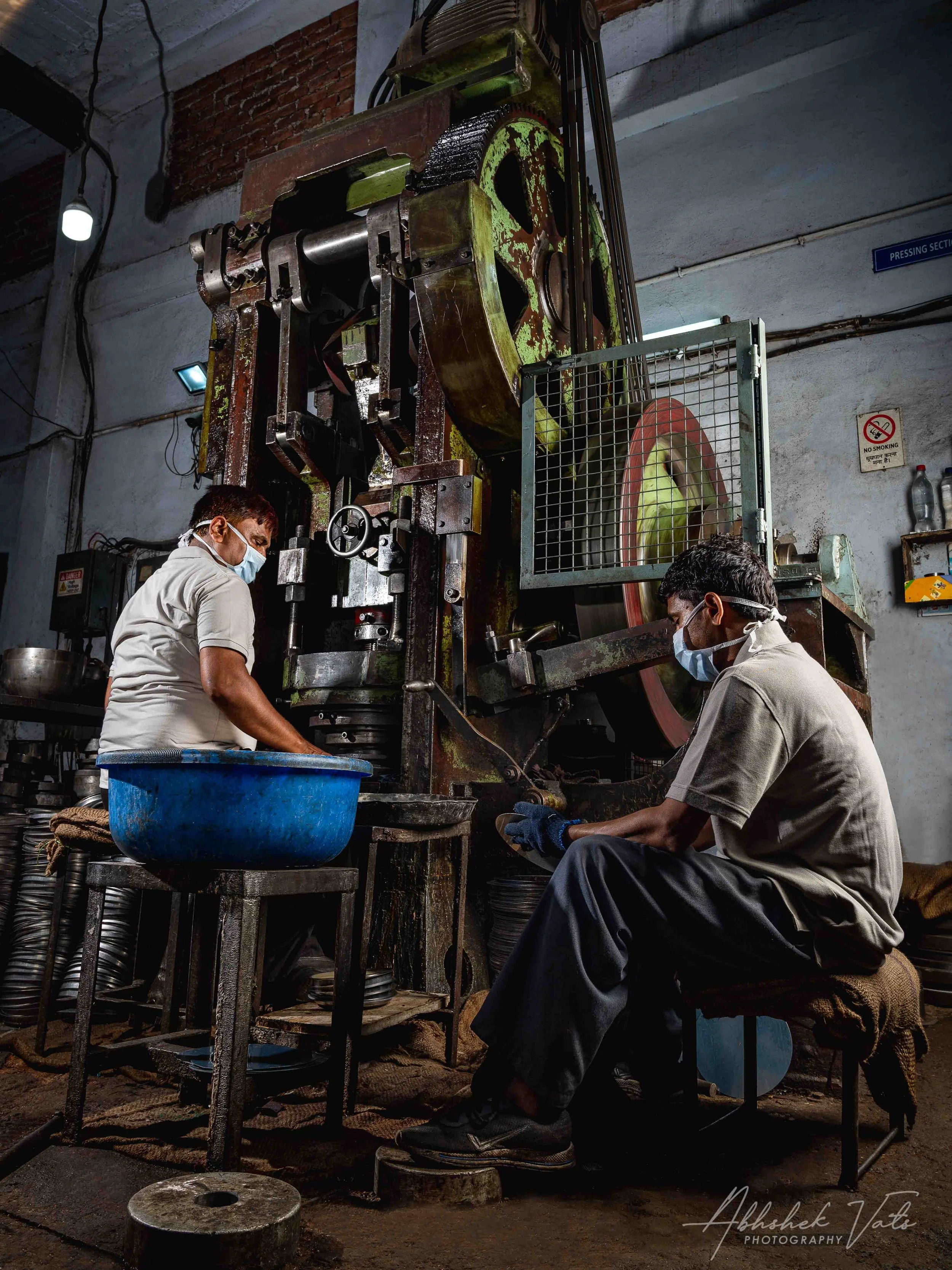 Two workers wearing face masks working on large industrial machinery in a workshop with concrete and brick walls.