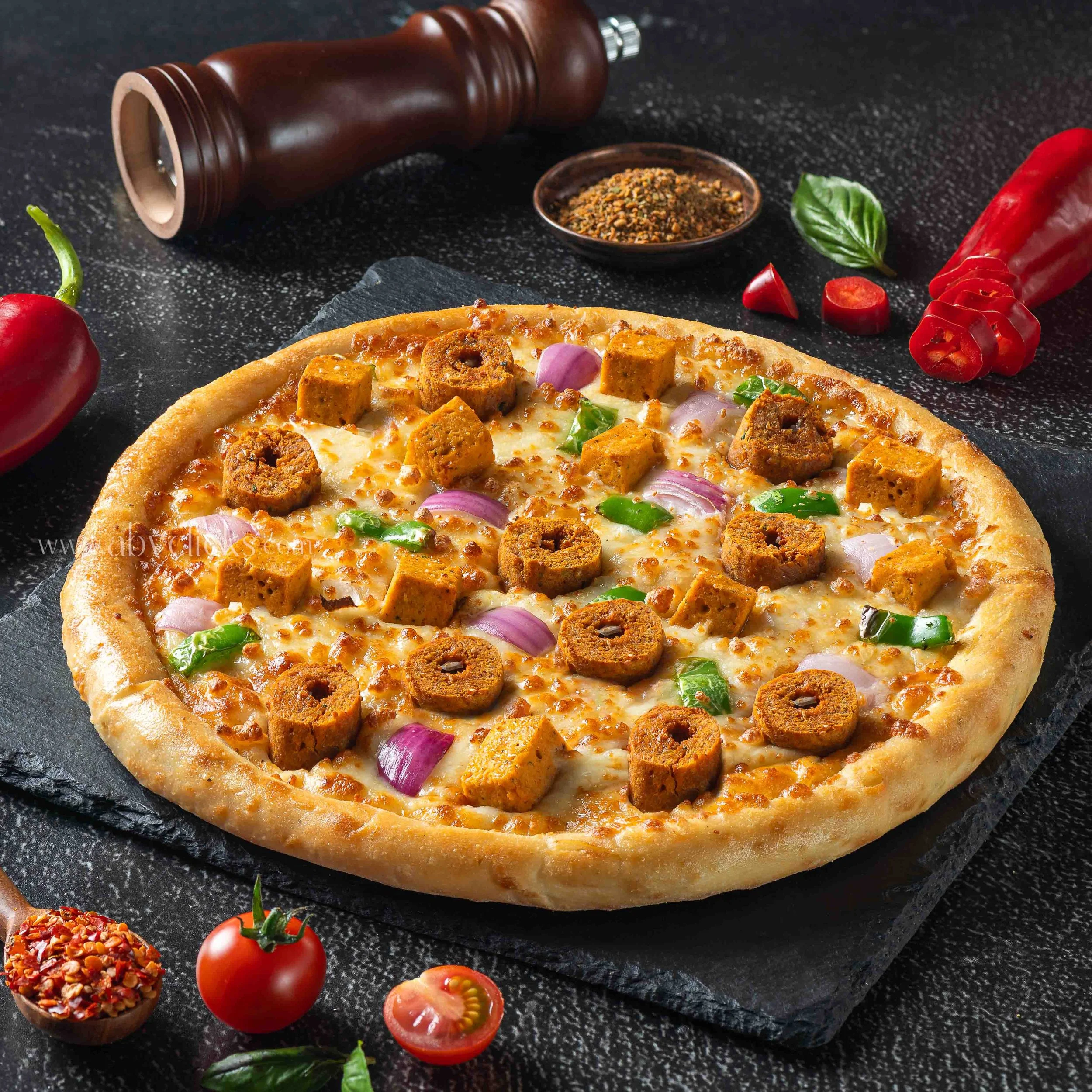 Close-up of a pizza with cheese, sausage, and vegetables on a dark surface. Surrounding are cherry tomatoes, a green chili pepper, fresh basil leaves, sliced cherry tomatoes, a bowl of crushed spices, and a pepper grinder.