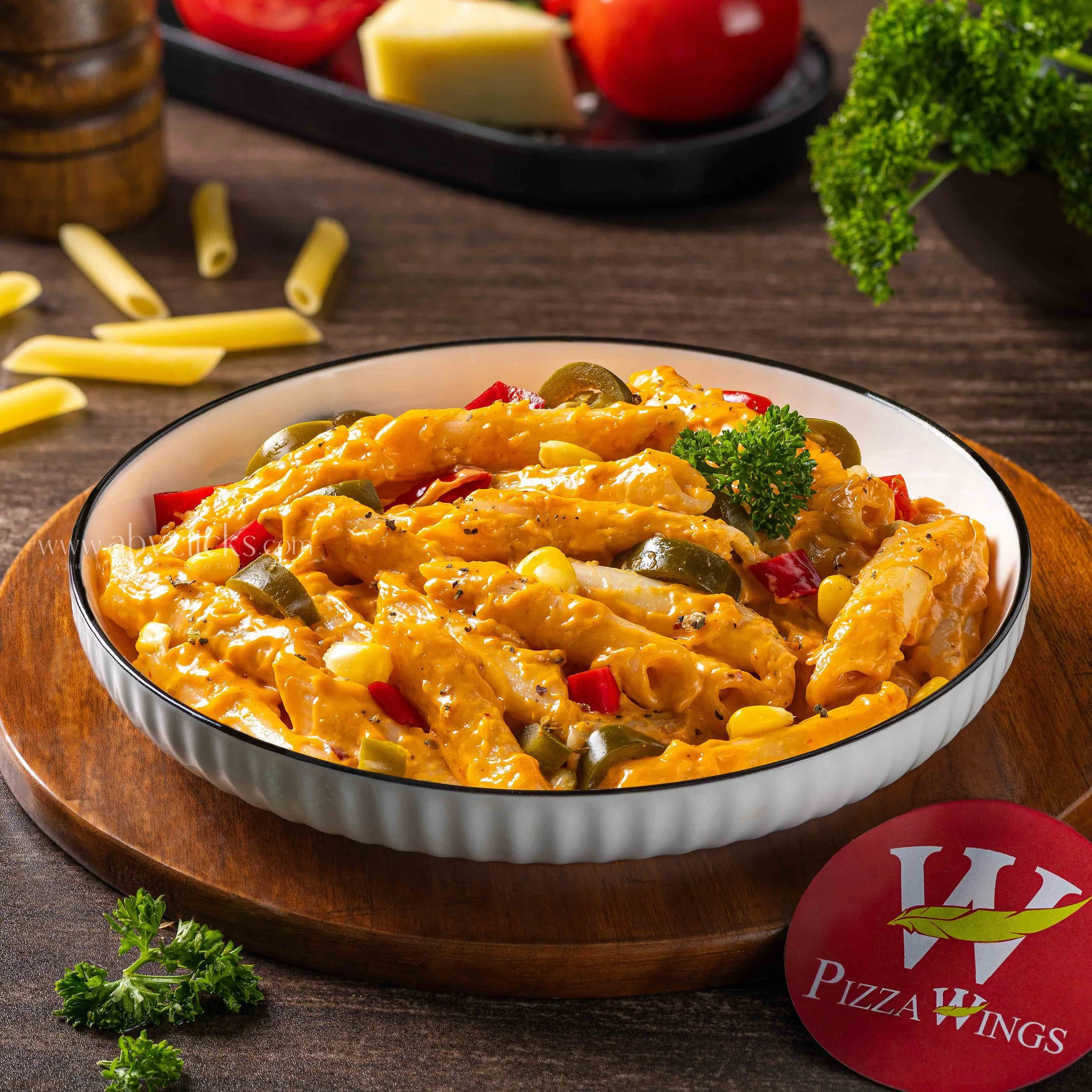 Close-up of a bowl of chicken and vegetable casserole garnished with parsley, with pasta, cheese, cherry tomatoes, and fresh herbs in the background.