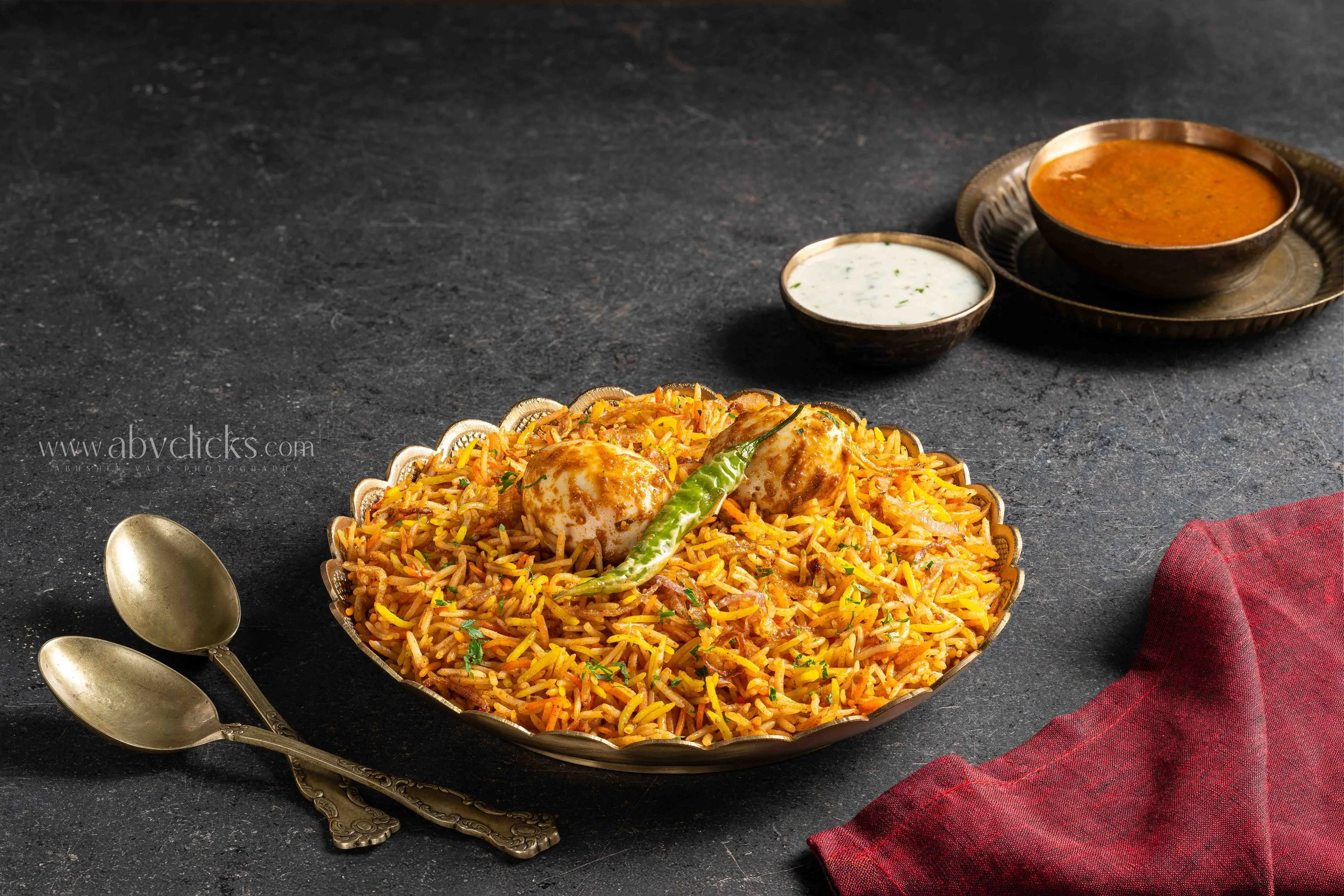 A large silver tray of spiced lemon rice garnished with herbs and a green chili, alongside three antique silver spoons on a dark textured surface, with small bowls of curry and yogurt in the background, and a red cloth napkin to the right.
