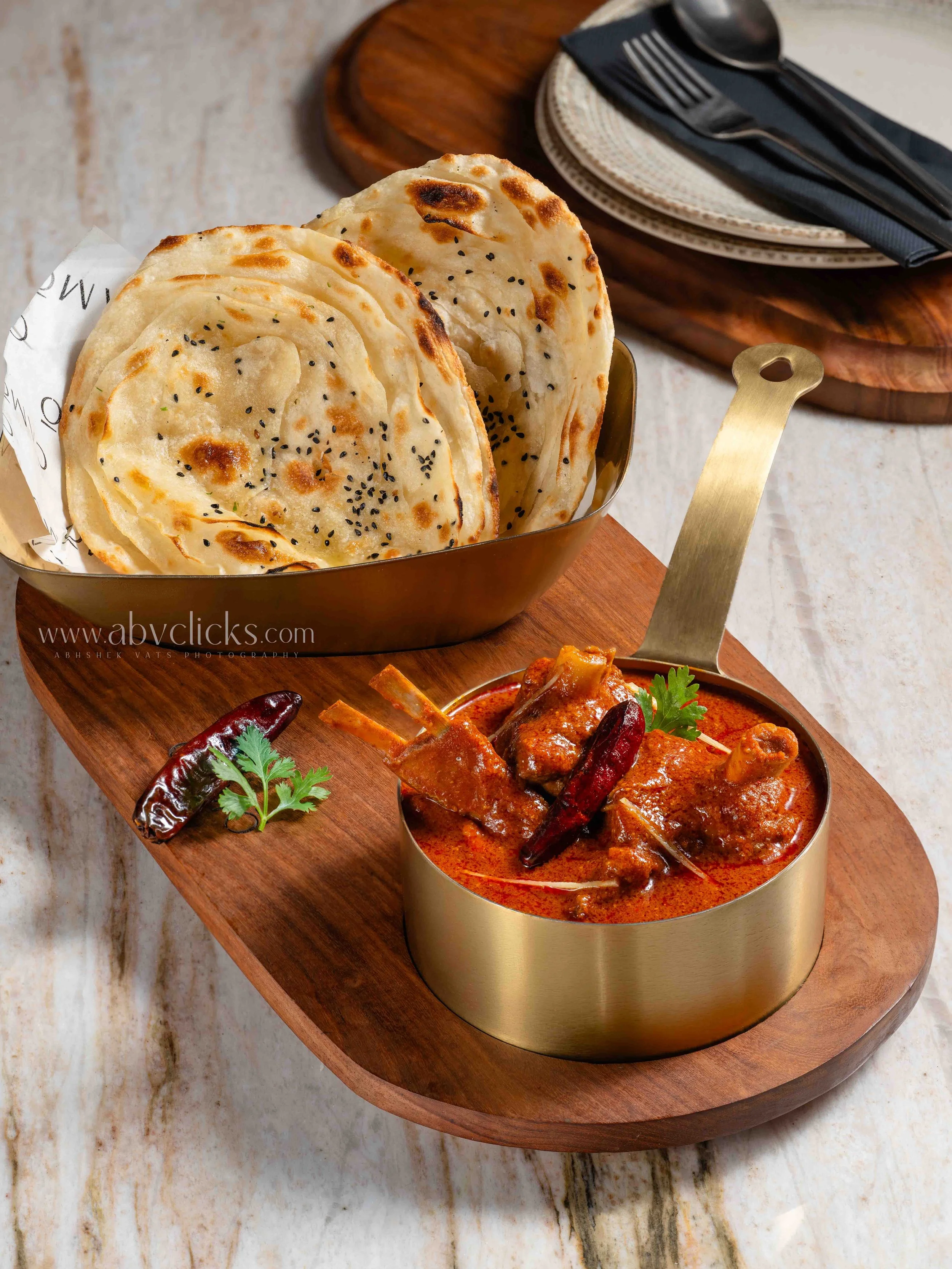 Stacked naan bread with black sesame seeds in a metal bowl on a wooden serving board, with a spicy Indian curry in another bowl garnished with cilantro and dried red chilies.