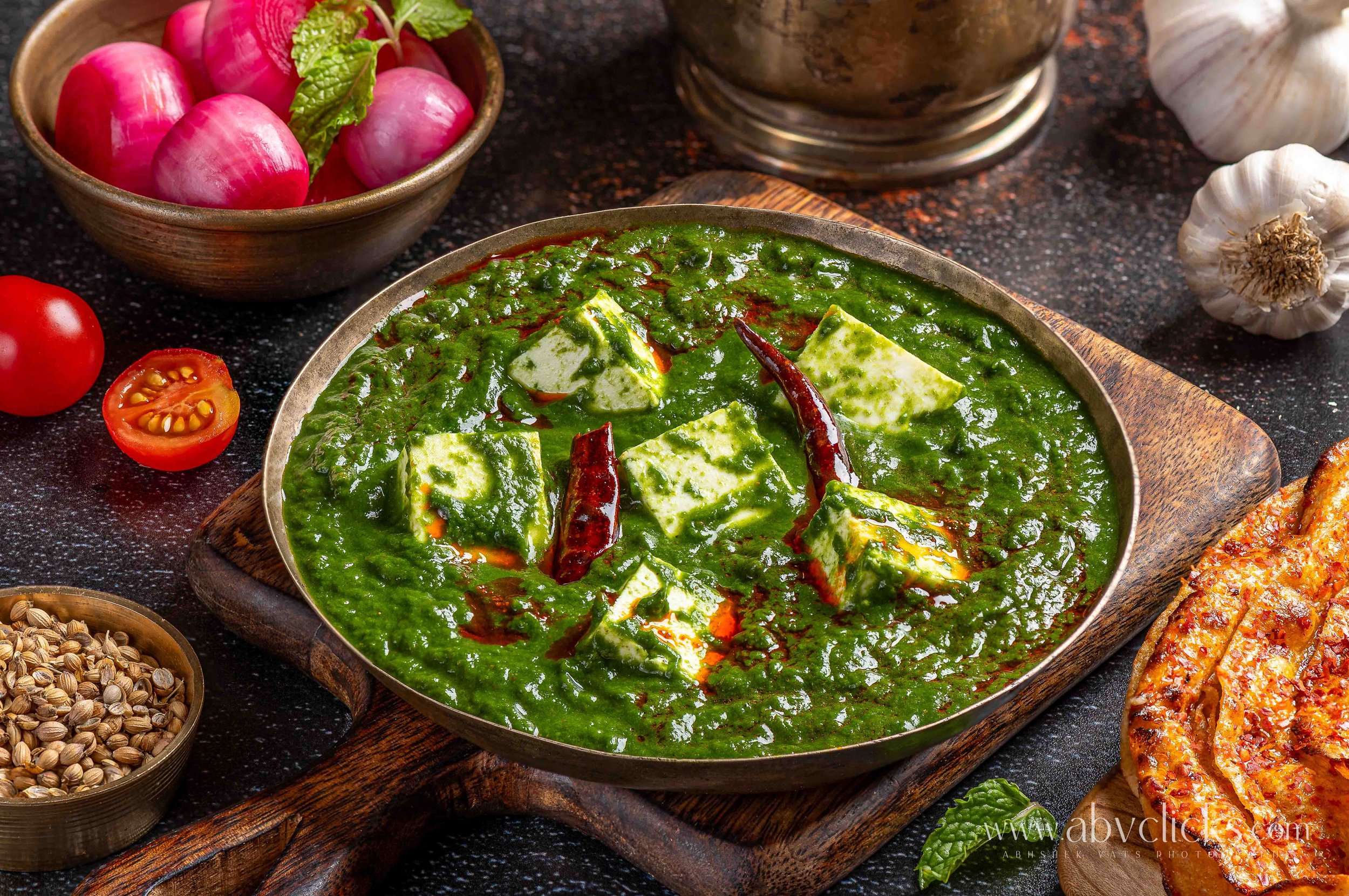 A bowl of green curry with tofu, dried red chilies, and herbs surrounded by small bowls of spices, radishes, tomatoes, garlic, and a piece of grilled meat on a dark textured surface.
