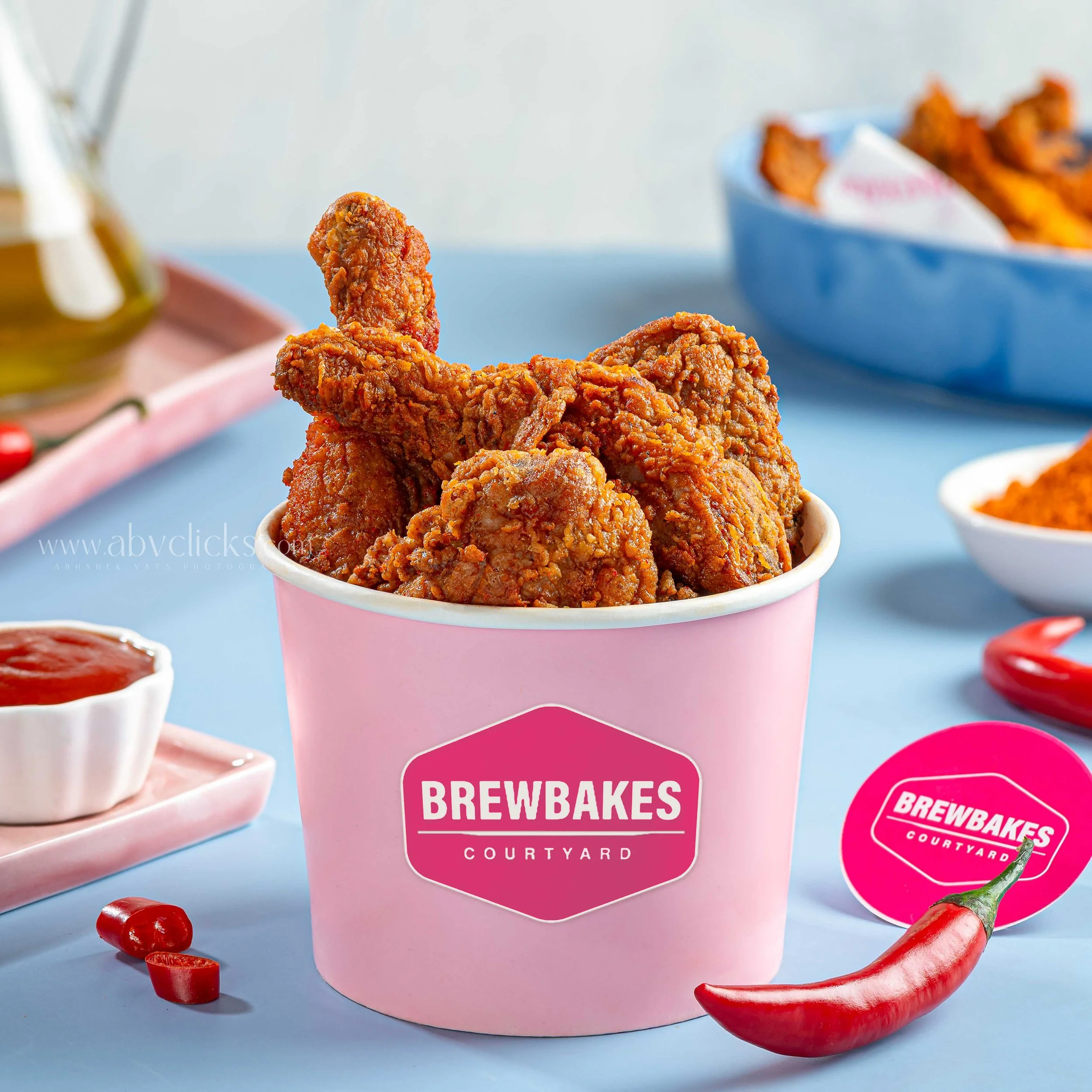 Close-up of a paper cup filled with crispy fried chicken pieces, with a pink container labeled "Brewbakes Courtyard" in the background and scattered red chili peppers on a light blue surface.