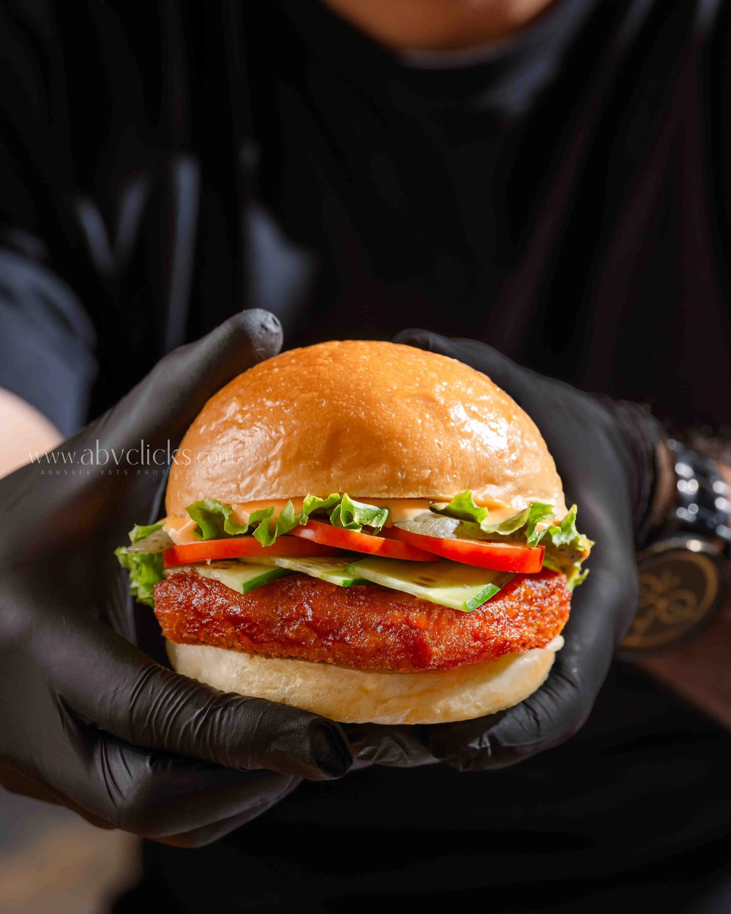 Close-up of a chicken sandwich held in black-gloved hands, featuring a golden bun, crispy fried chicken fillet, lettuce, tomato, and mayonnaise.