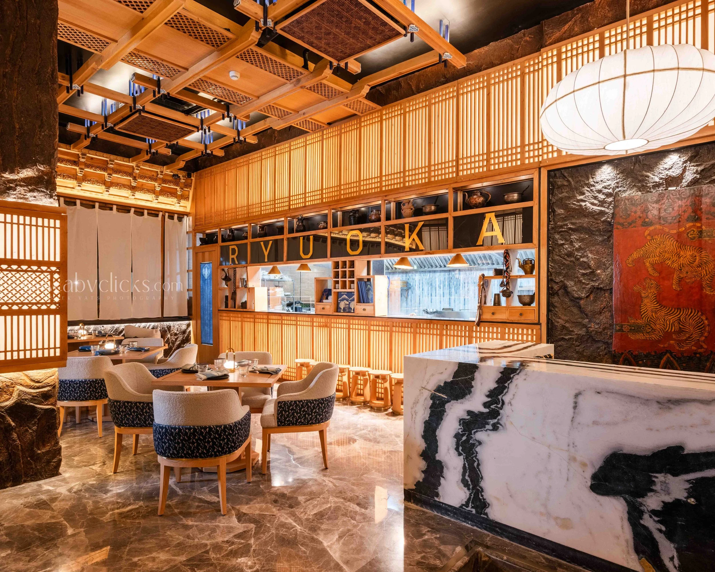 Interior of a restaurant with wooden lattice walls, marble countertops, and a ceiling with wooden beams and black panels. There are tables with chairs, decorative items, and a lantern-style light fixture.