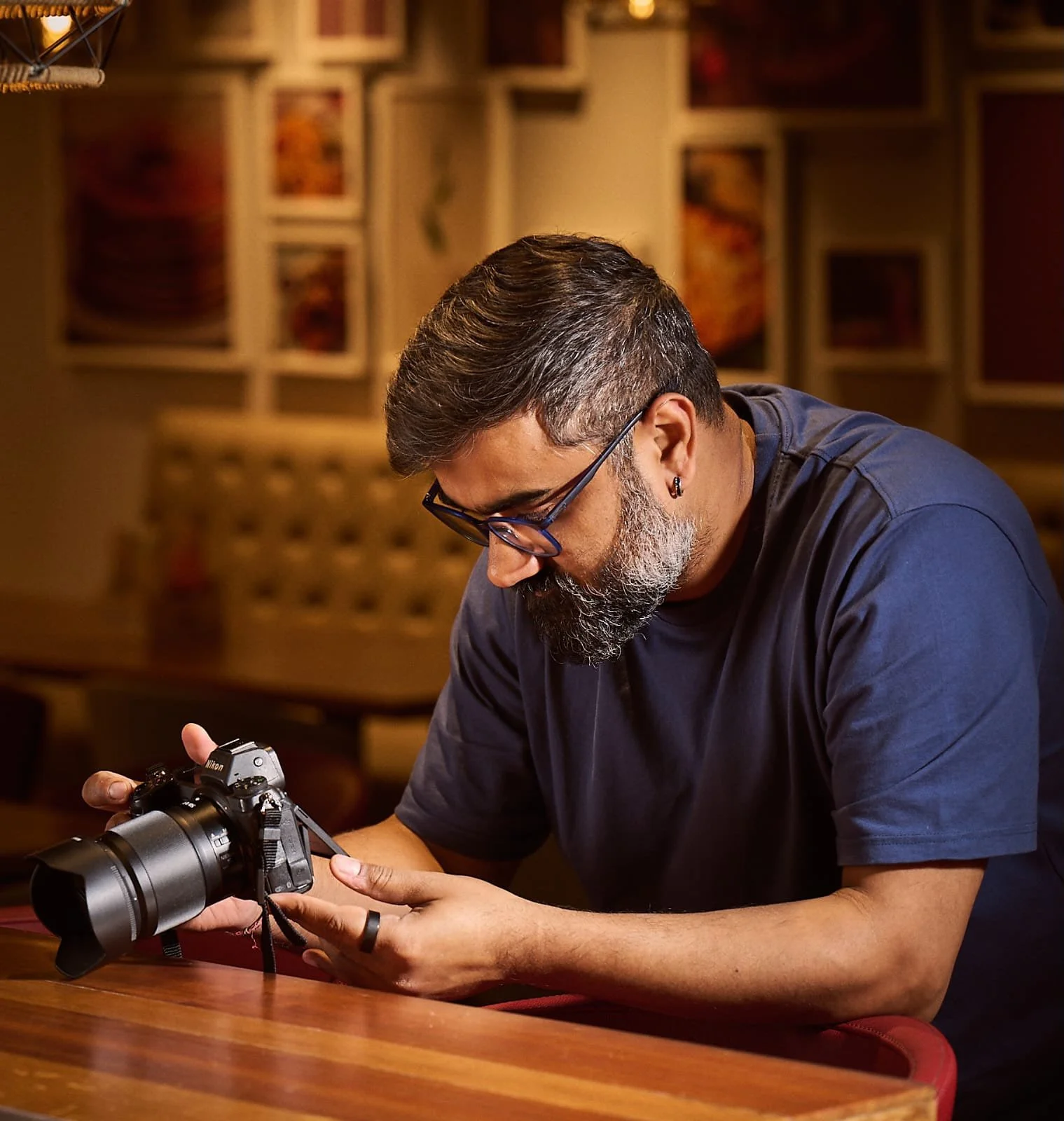A man with glasses and a beard, wearing a dark blue shirt, is sitting at a wooden table examining a camera. The background features framed pictures on the wall.