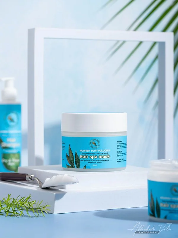 A hair spa mask container on a white tray with a spa brush, a green leafy branch, and other skincare products in the background, set against a light blue wall.