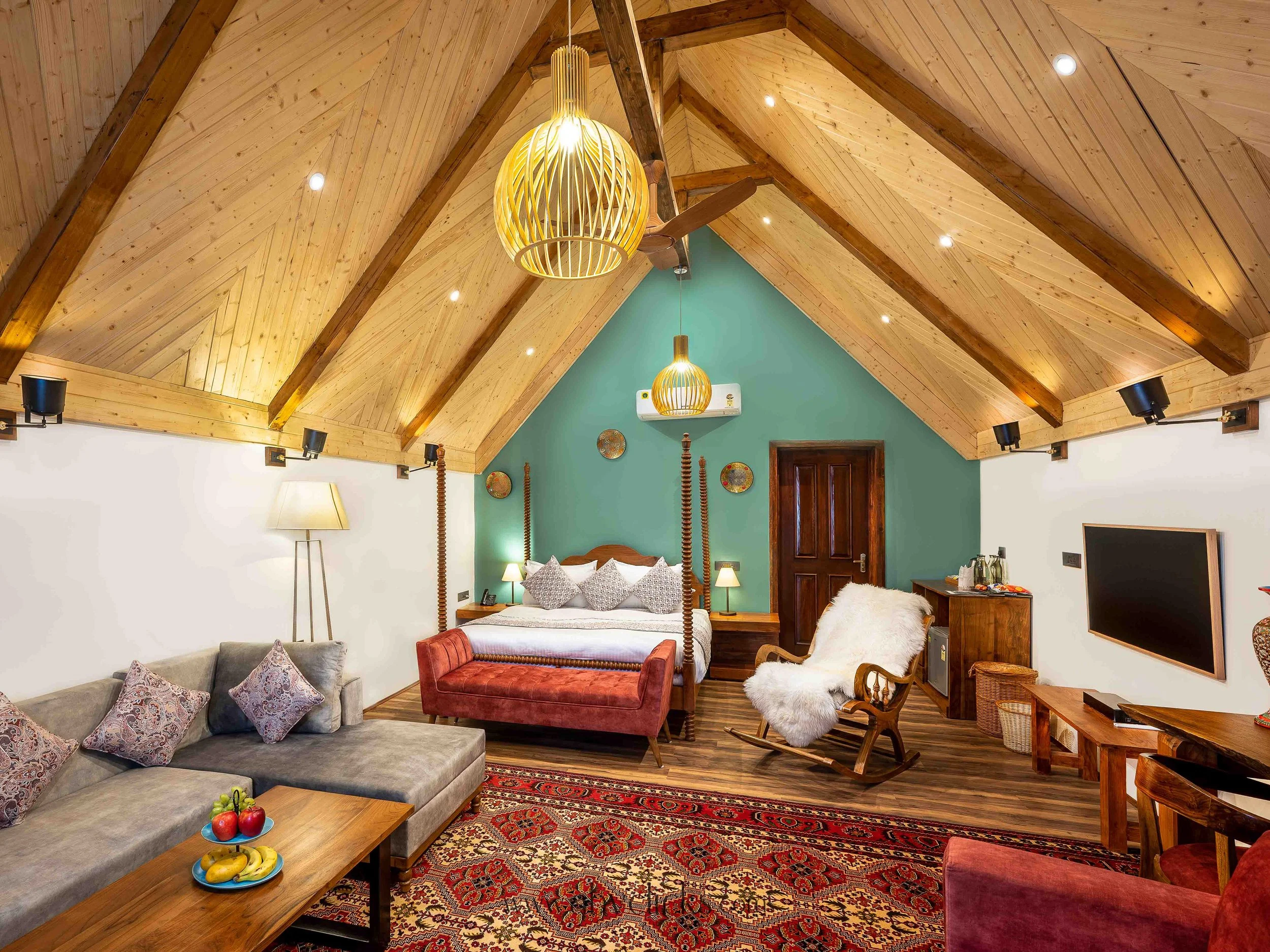For brand Rahbag by the Orchard, Srinagar, Interior of a cozy room with a wooden vaulted ceiling, two pendant lights, a four-poster bed, a gray sofa with patterned pillows, a wooden coffee table with apples and bananas, and a colorful Persian rug.