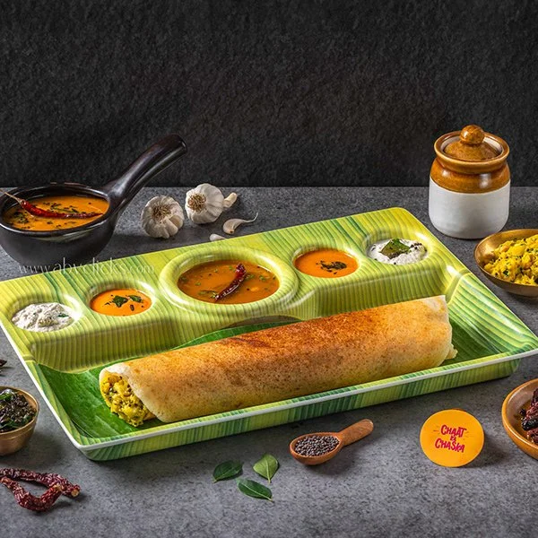 Plate with a rolled Indian-style stuffed pancake and multiple small bowls of colorful sauces and chutneys on a green tray, with garlic, spices, and condiments around.