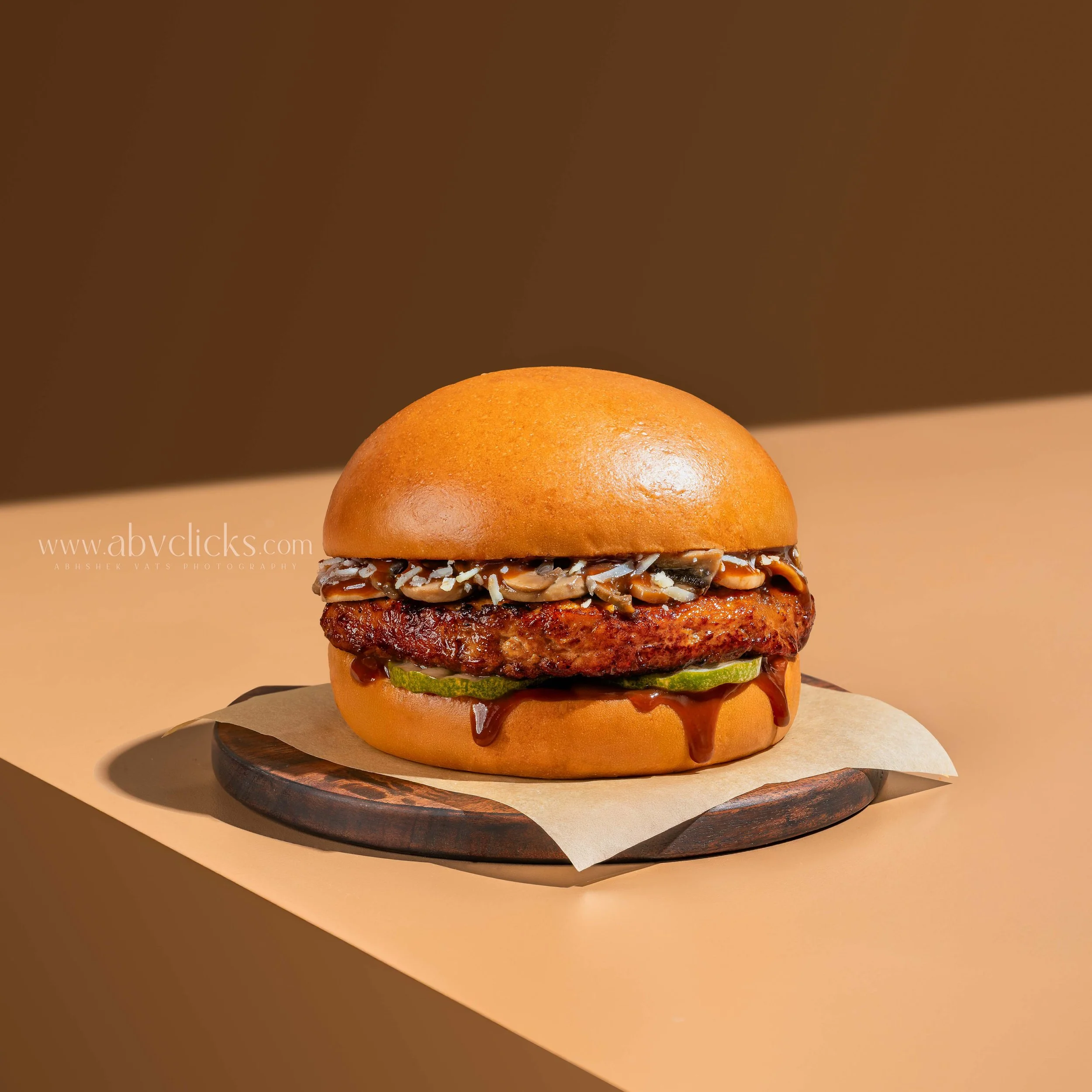 Close-up of a gourmet burger with a toasted bun, lettuce, grilled patty, caramelized onions, sliced pickles, and a drizzle of sauce, on a round wooden serving board against a brown background.