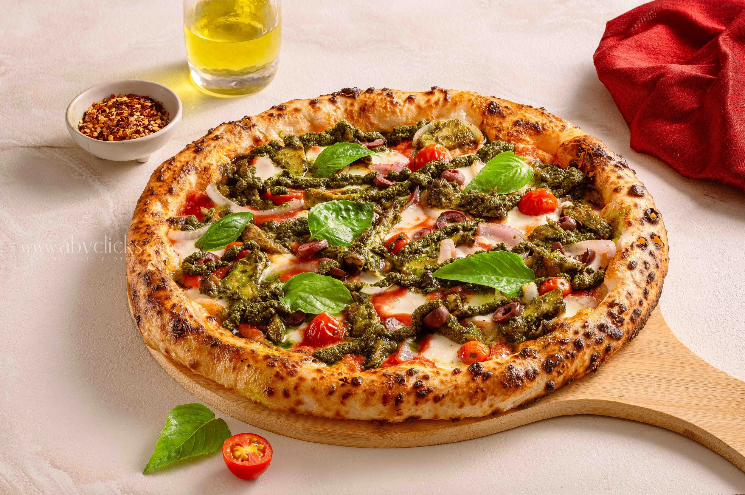 A pizza with a golden crust, topped with green pesto, cherry tomatoes, sliced onions, black olives, melted cheese, and fresh basil leaves, on a round wooden board with a glass of yellow beverage and a small bowl of crushed red pepper flakes nearby.