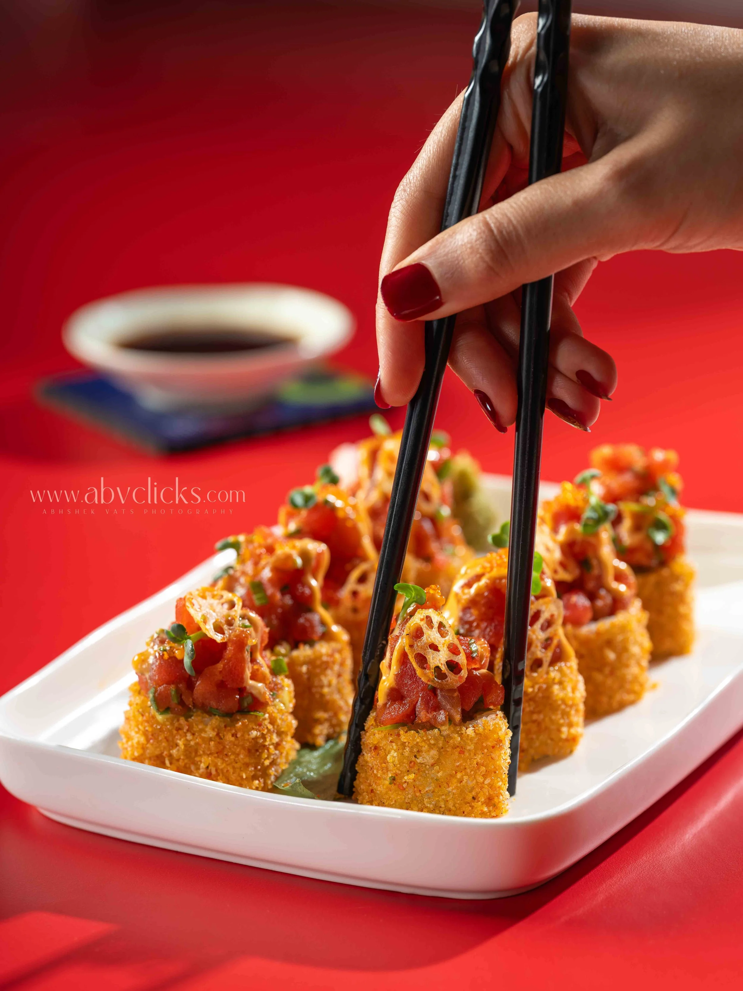 Close-up of a hand using black chopsticks to pick up pieces of crispy fried sushi roll topped with spicy sauce and garnished with chopped green onions on a white plate, with a small cup of soy sauce in the background on a red table.