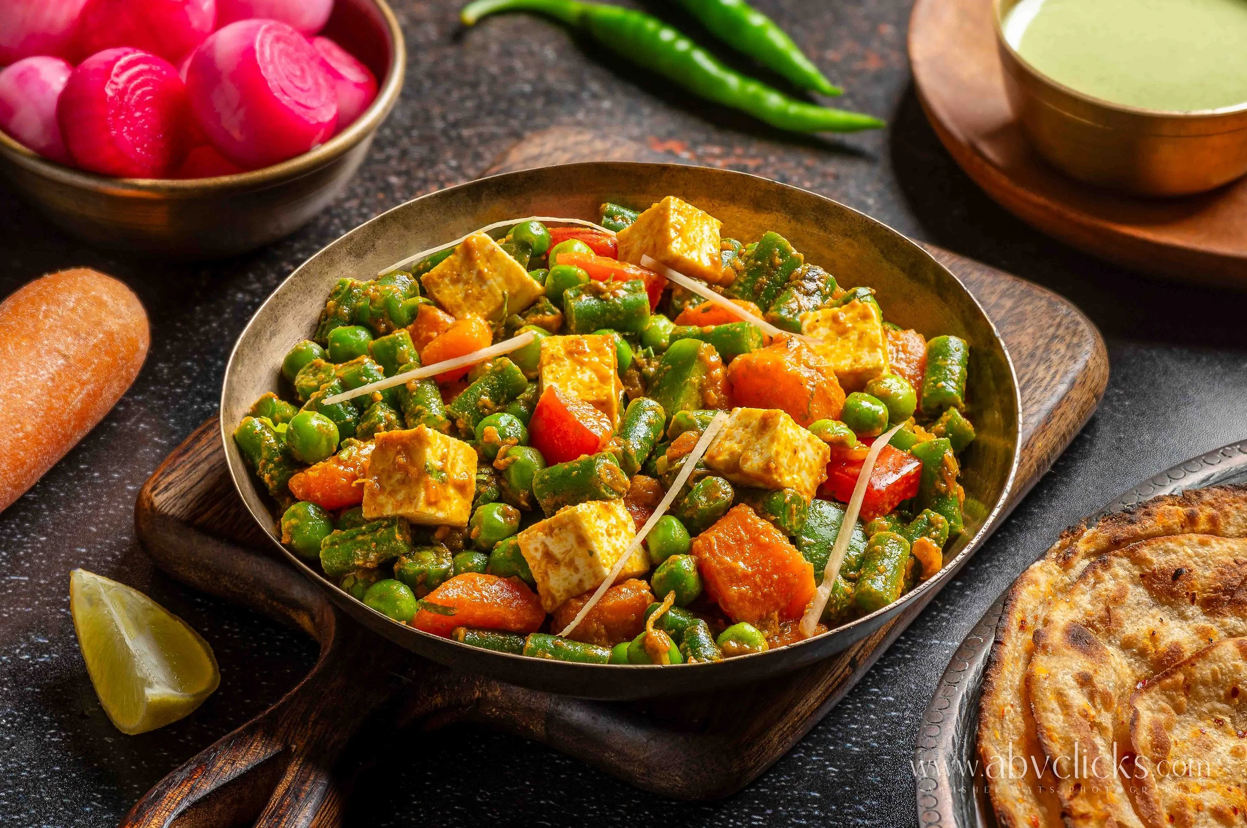 A dish of Indian vegetable curry with peas, carrots, and paneer cheese served in a black bowl, accompanied by lime wedges on a dark surface with other bowls and plates of food and green chili peppers in the background.