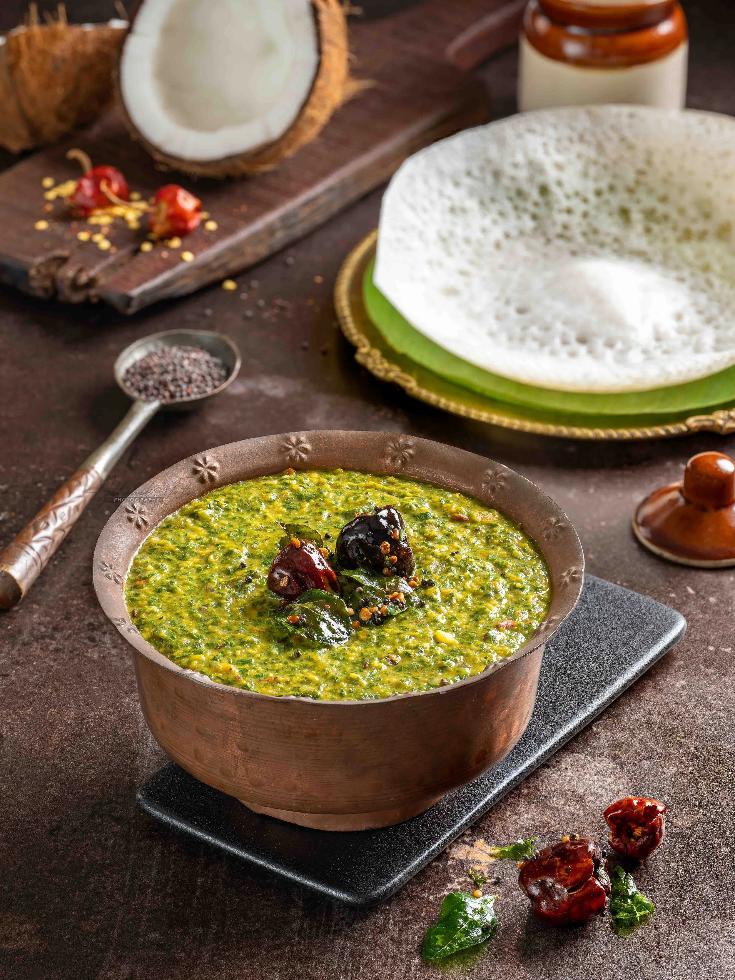 A bowl of cooked spinach or a similar green leafy vegetable dish garnished with black and red spices and dried berries, placed on a black stand on a rustic dark brown surface, with scattered dried red chilies and green leaves around.