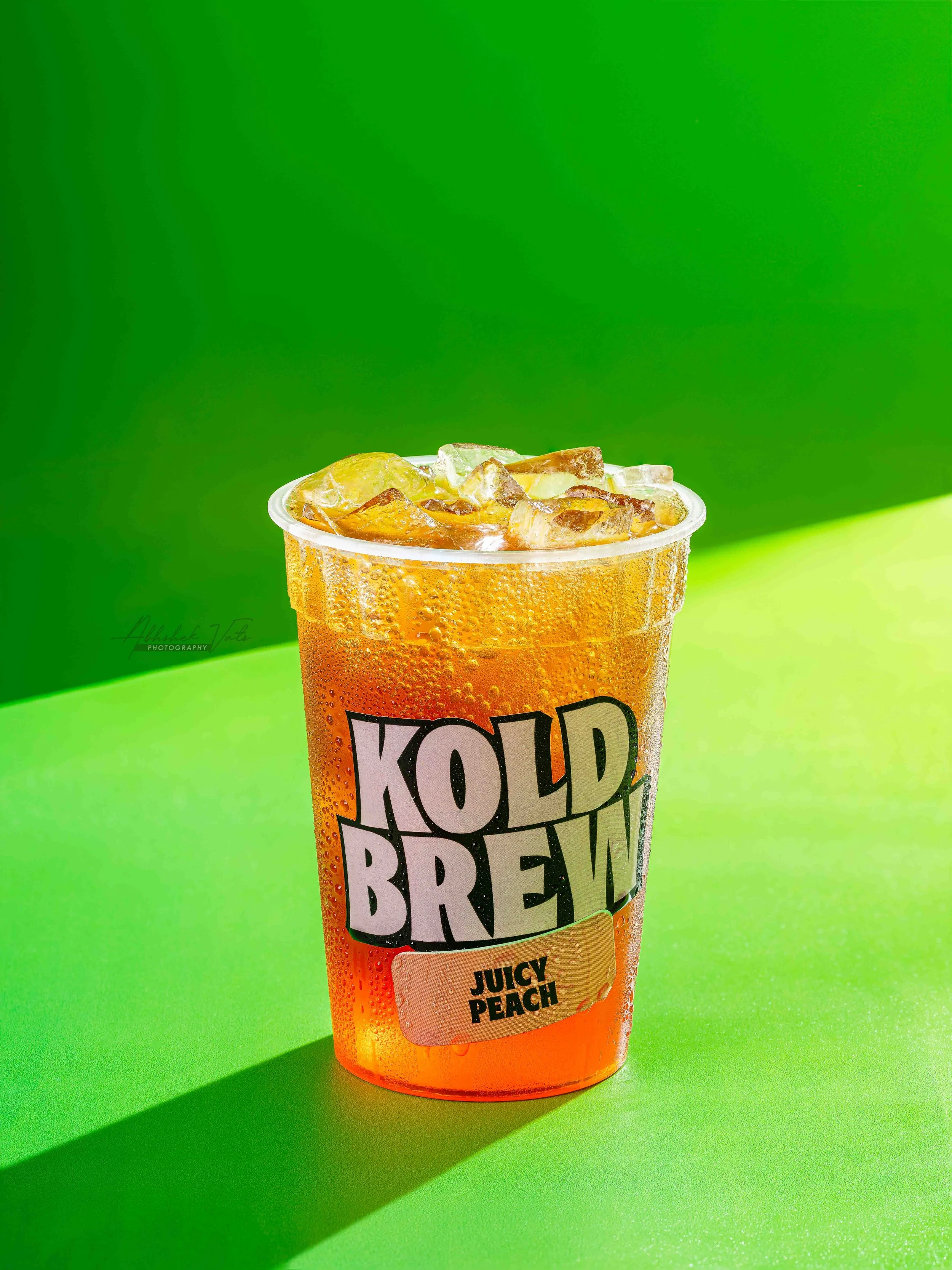 A plastic cup filled with an orange-colored carbonated beverage, ice cubes, and a lemon wedge, placed on a green surface with a bright green background.
