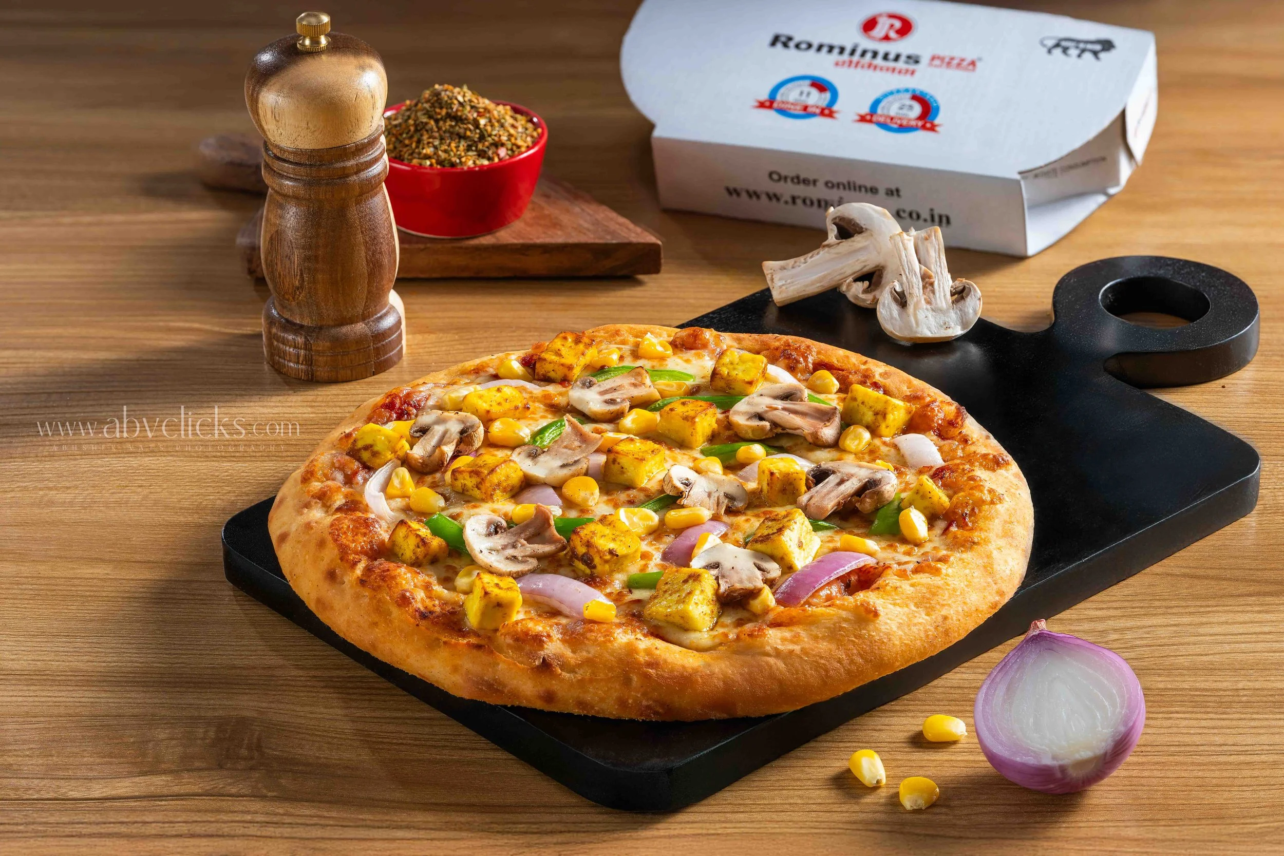 A pepperoni pizza with mushrooms, green bell peppers, yellow corn, and onions on a black pizza tray. Surrounding the pizza are sliced onions and a black pizza cutter. In the background are a wooden pepper mill, a red bowl of mixed herbs, a pizza box,