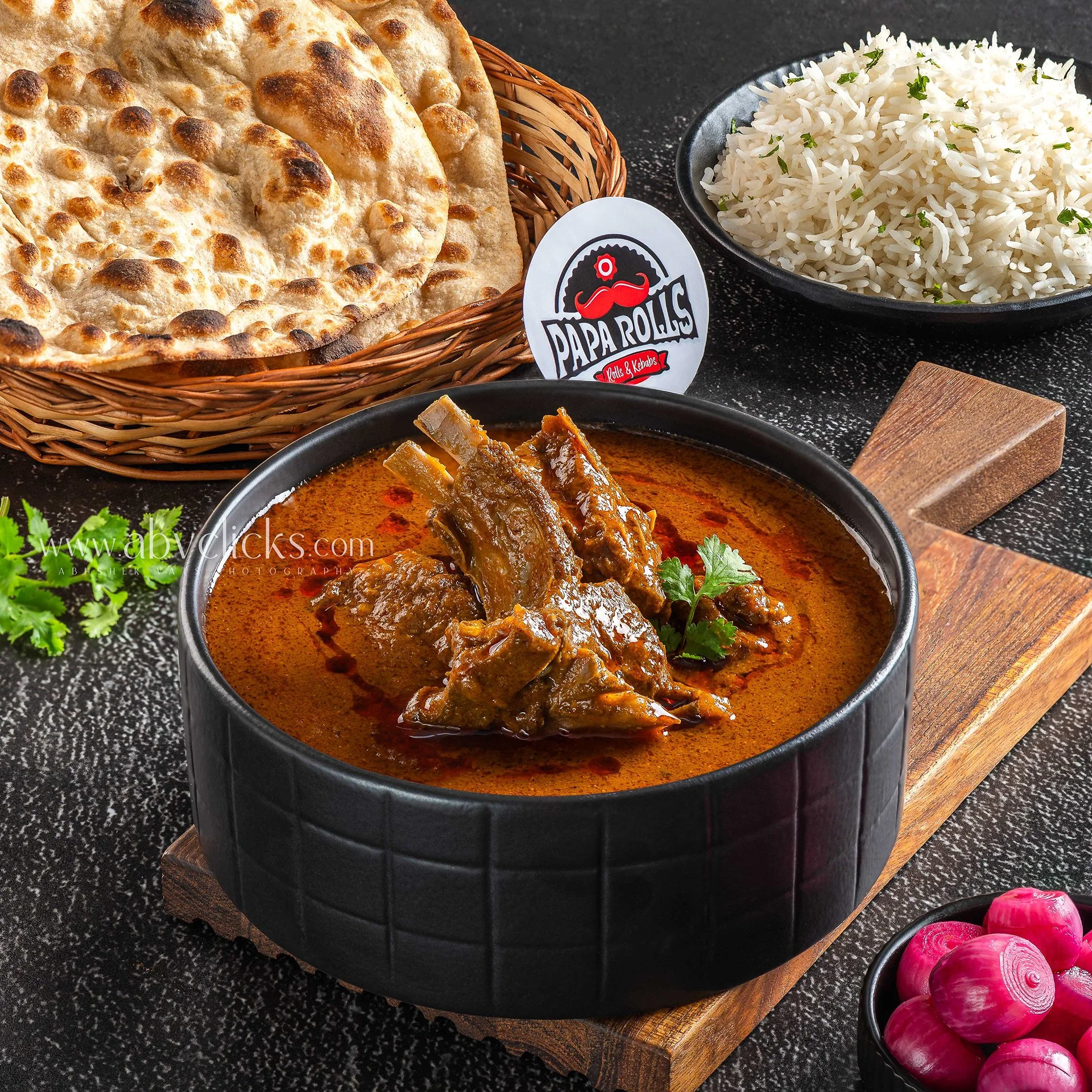 Indian cuisine with naan bread, rice garnished with herbs, and a bowl of lamb curry on a black surface with a wooden cutting board and a side bowl of sliced radishes.