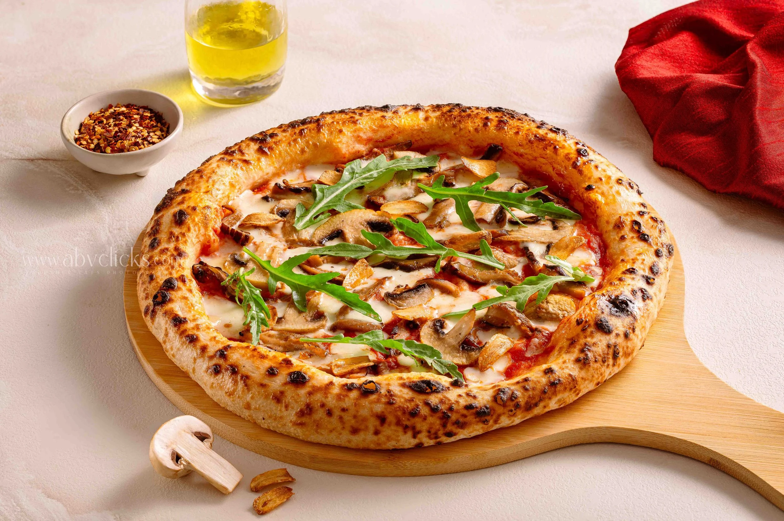 Cheese pizza with mushrooms, arugula, and garlic slices on a wooden pizza board, with a side of red pepper flakes, a glass of yellow beverage, and sliced mushrooms.