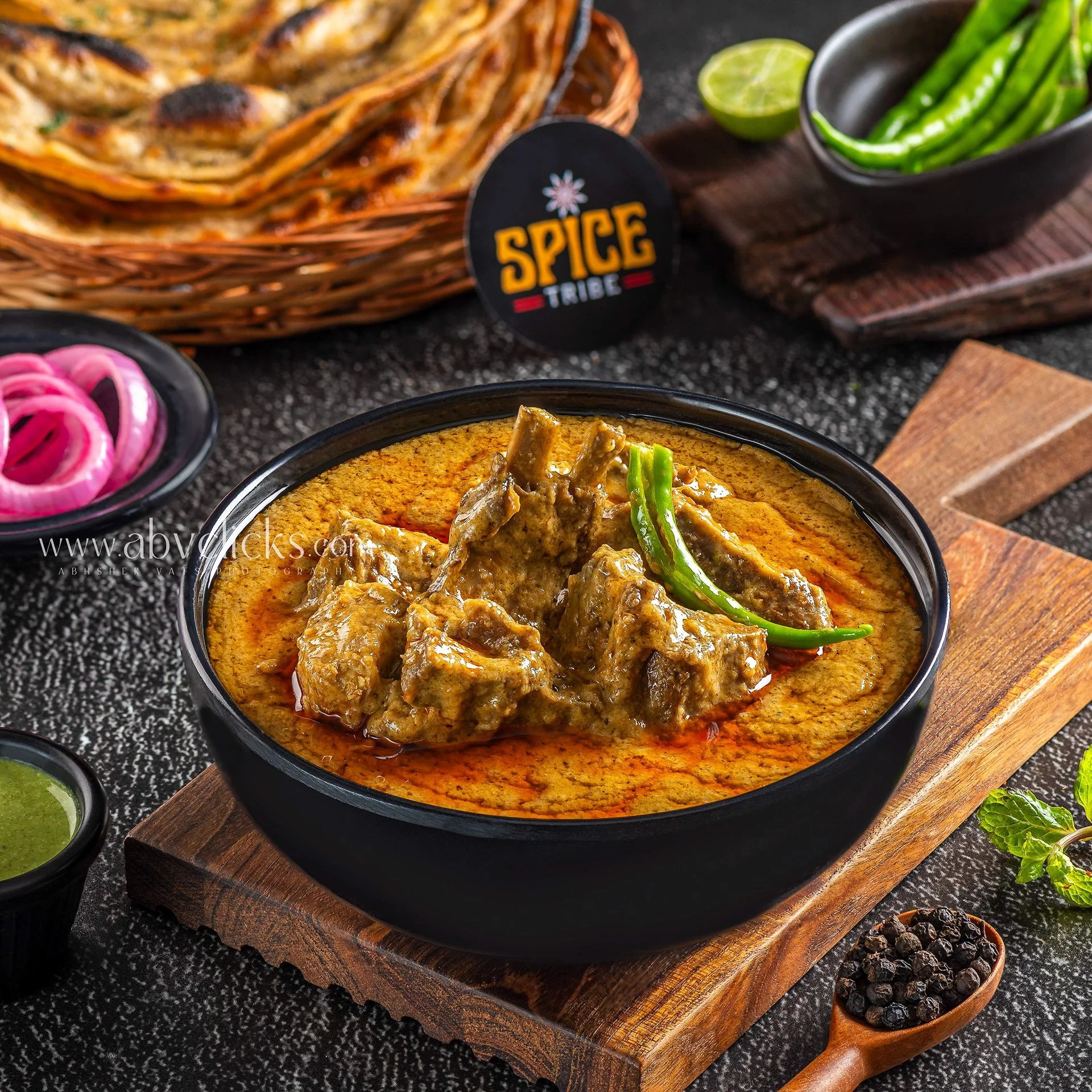 A bowl of lamb curry served on a wooden platter, garnished with green chilies. In the background, there are green chilies, a sliced lime, and pink pickled onions. A basket of flatbread and small bowls of green sauce and black pepper are also visible.