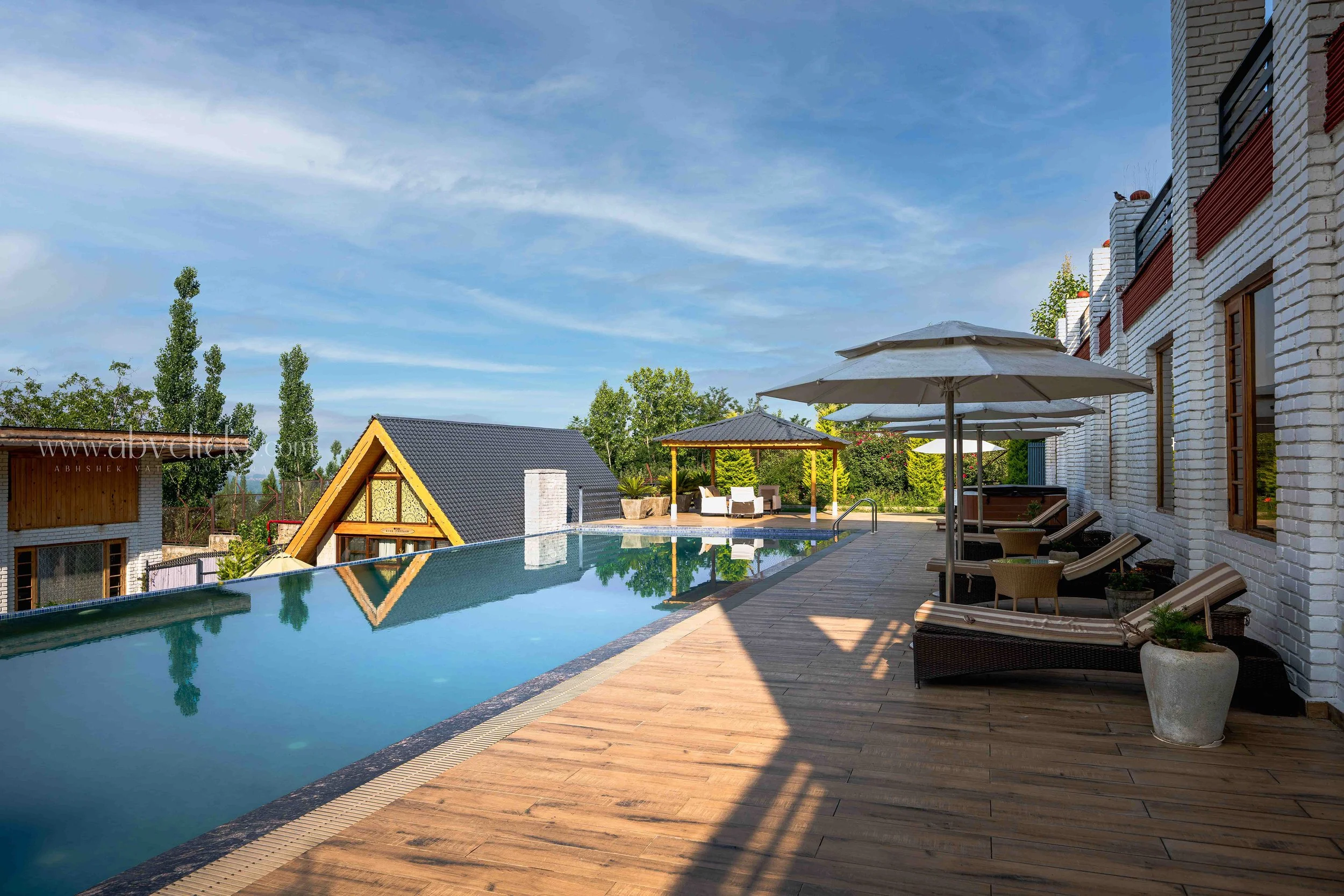 A backyard with a swimming pool, wooden deck, white brick house, patio chairs, and umbrellas, with a small gazebo, trees, and houses in the background on a clear day.
