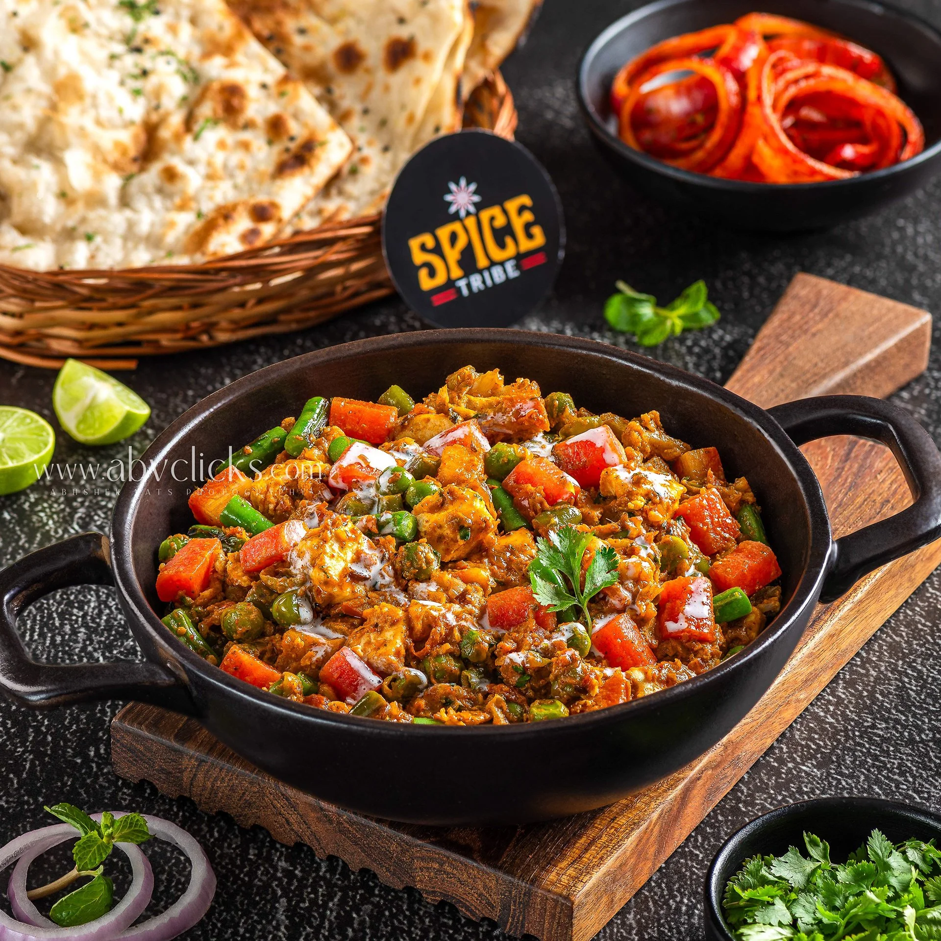 A black bowl of vegetable curry garnished with cilantro, on a wooden board, with lime wedges and sliced onions with mint on the side.