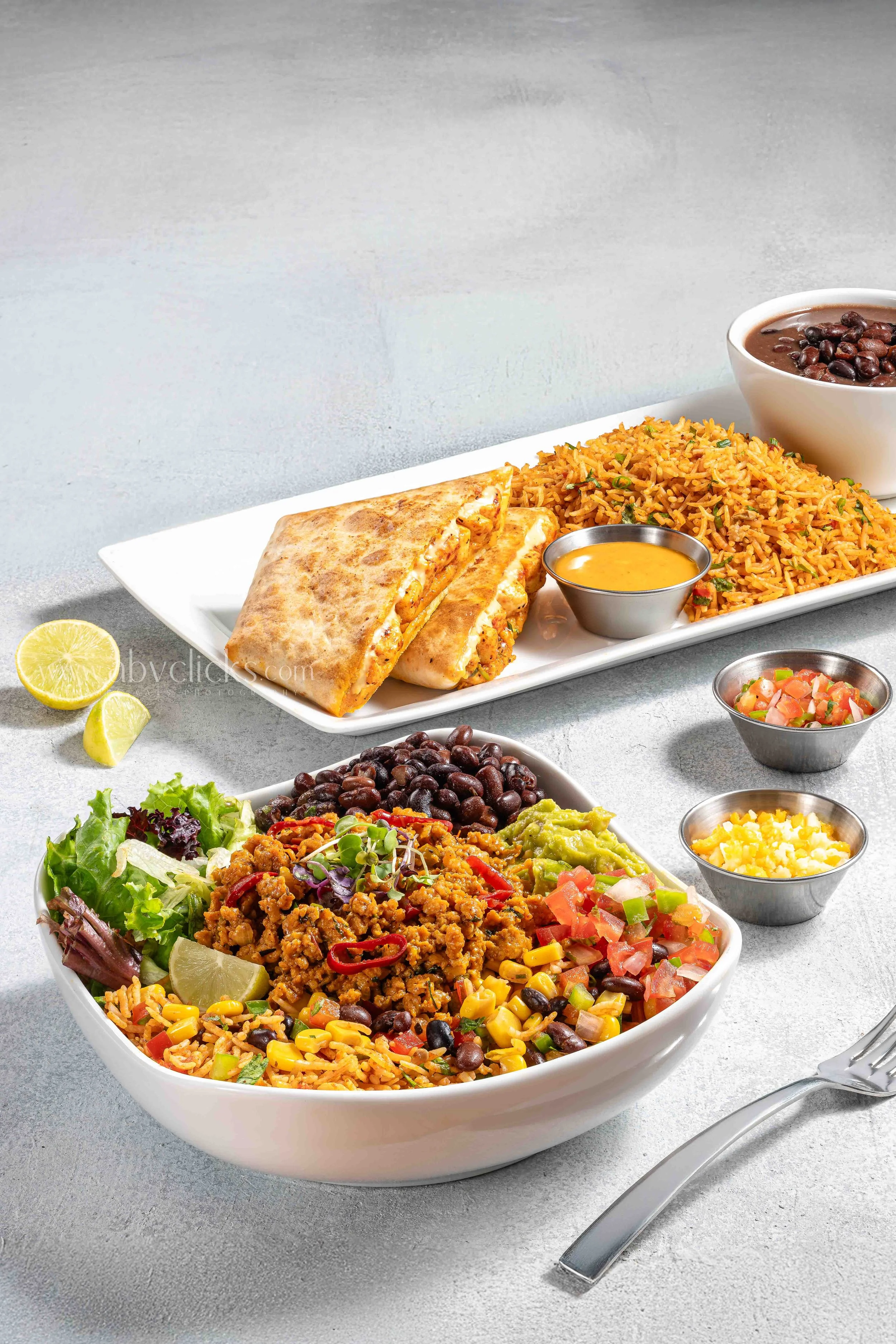 Colorful Mexican dishes including a large salad with corn, black beans, tomatoes, and ground meat, a bowl of rice, a quesadilla, and small containers of salsa, cheese, and sauce on a table with lime wedges and a fork.