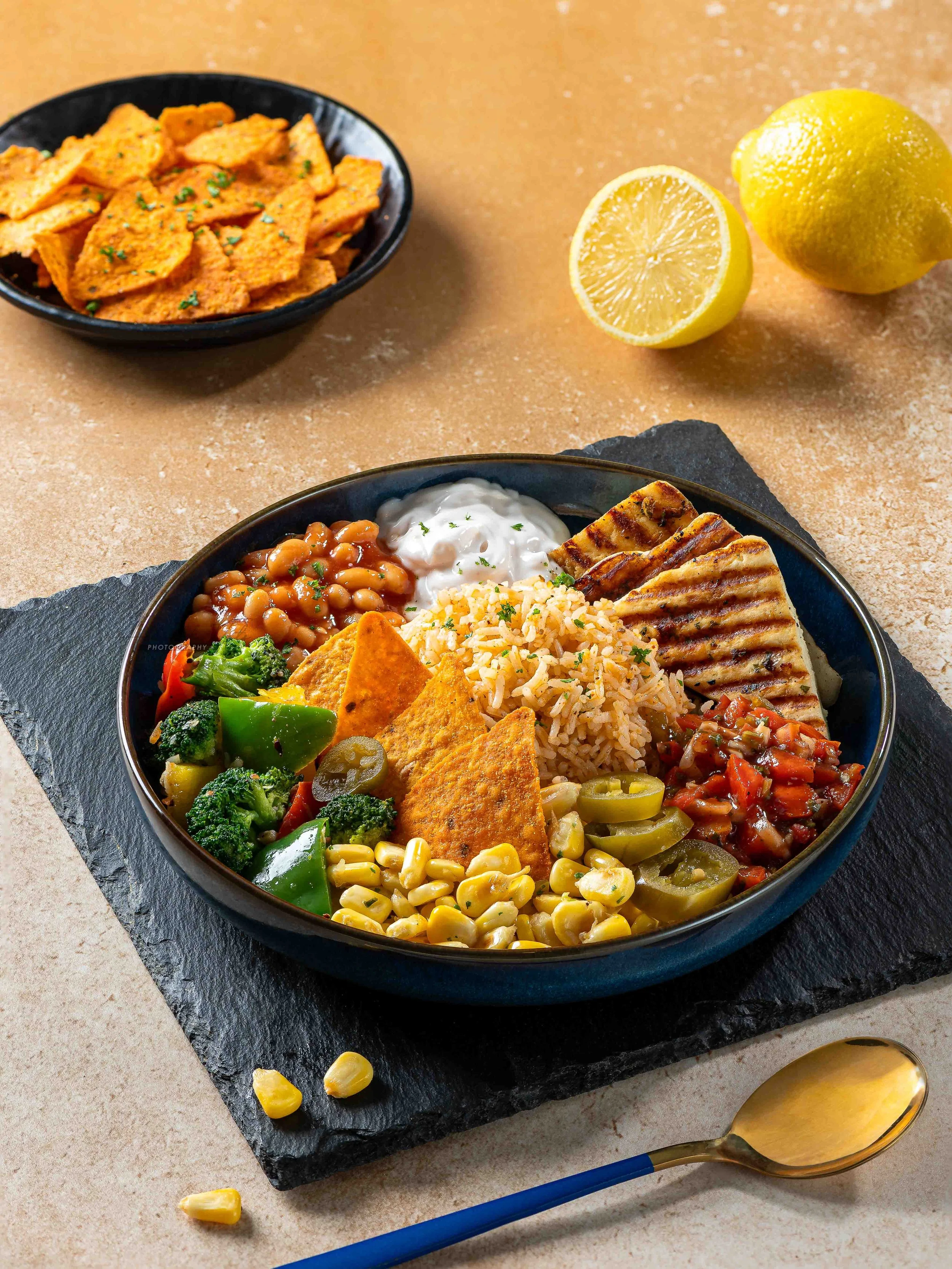 A round black bowl filled with Mexican food, including rice, beans, grilled chicken, vegetables, nachos, and sour cream, placed on a black slate serving board. There is a halved lemon and a whole lemon in the background on a beige surface, with a gol