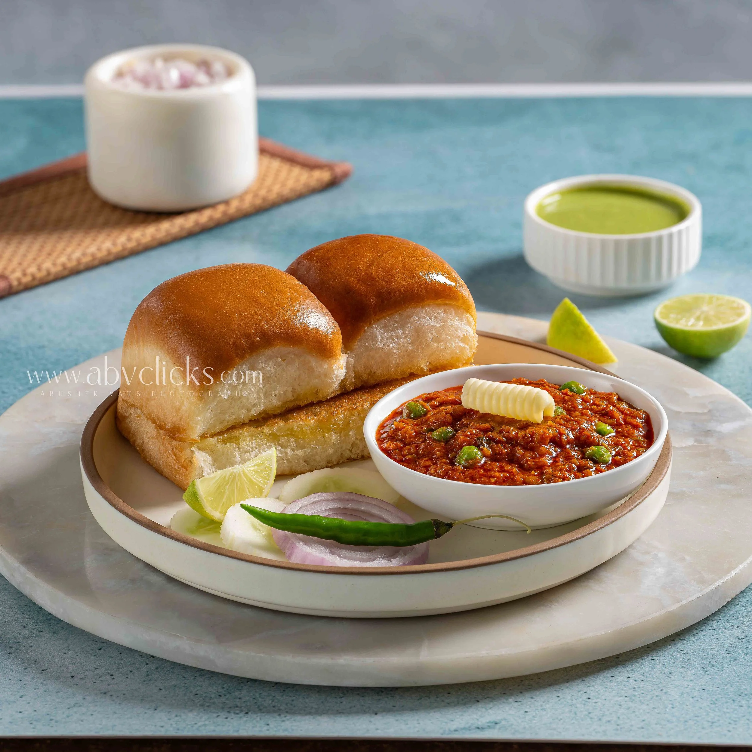 Close-up of a plate with two soft but crispy dinner rolls, garnished with lime wedges, sliced onions, and green chili peppers, with a bowl of spicy chutney or curry, on a textured surface.