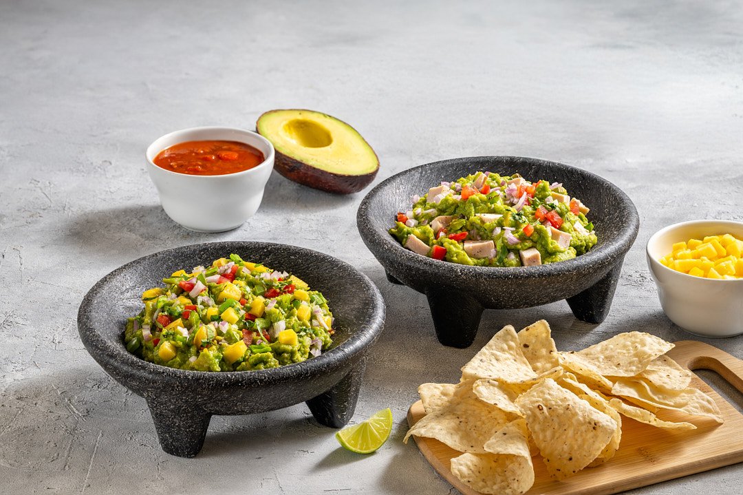 Two black stone bowls of chopped vegetable salads, one with diced chicken, along with a small bowl of shredded cheese, a bowl of salsa, a bowl of diced pineapple, sliced avocado, tortilla chips, and a lime wedge.
