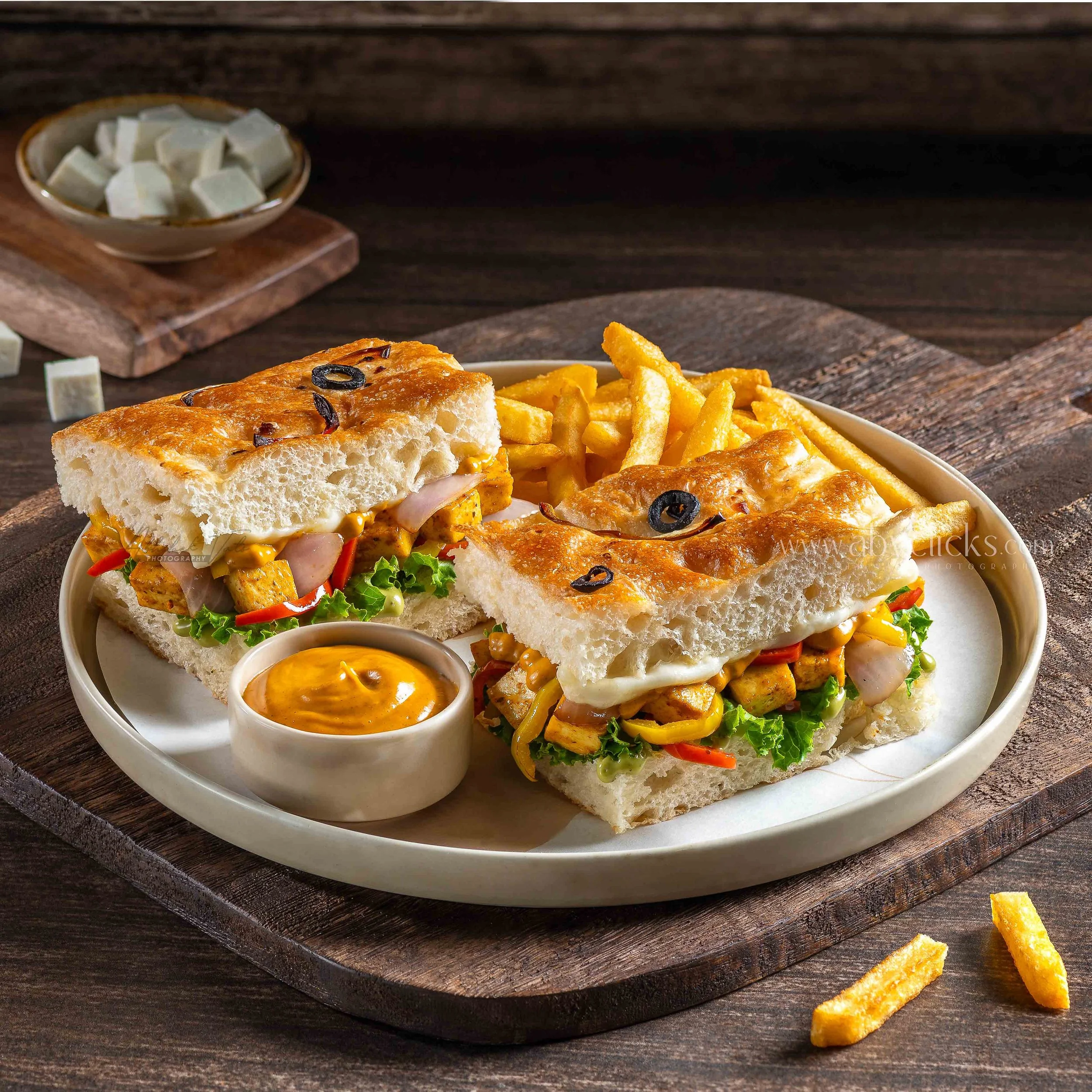 A plate with two slices of pizza topped with black olives, lettuce, tomatoes, and onions, alongside French fries and two small cups of dipping sauce, on a dark wooden surface with a piece of lemon on the side.