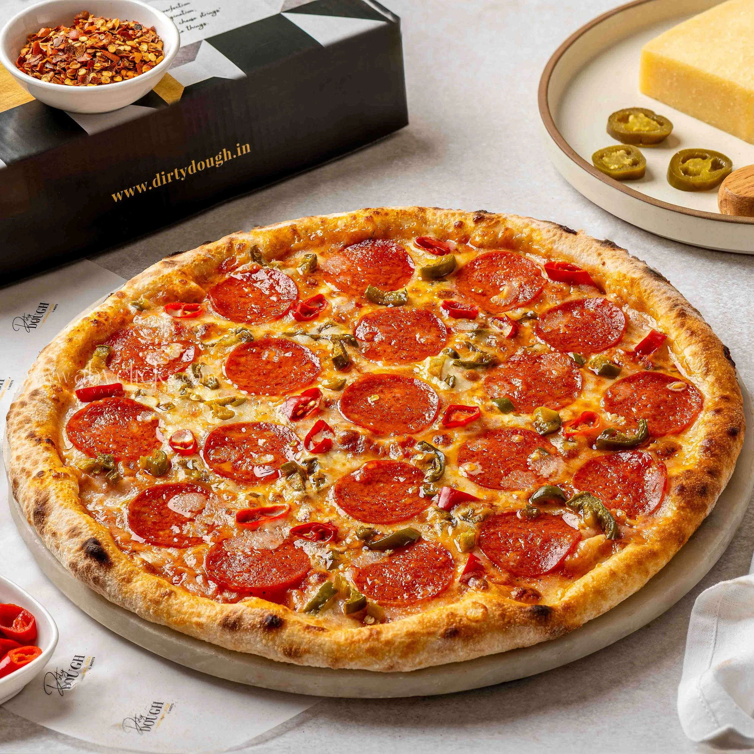 A pepperoni pizza with green peppers on a white plate, with a small bowl of sliced red chili peppers and a box of dried chili flakes in the background.