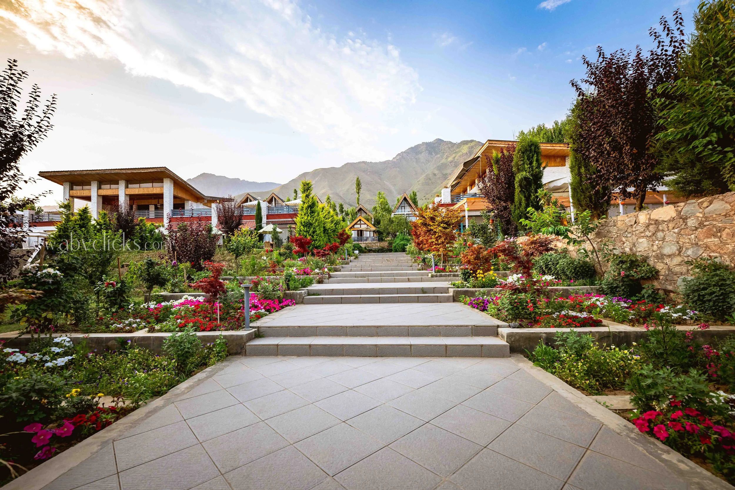 Stone pathway leading to colorful flower garden with various trees and plants, residential houses with modern architecture, mountains and blue sky in the background.