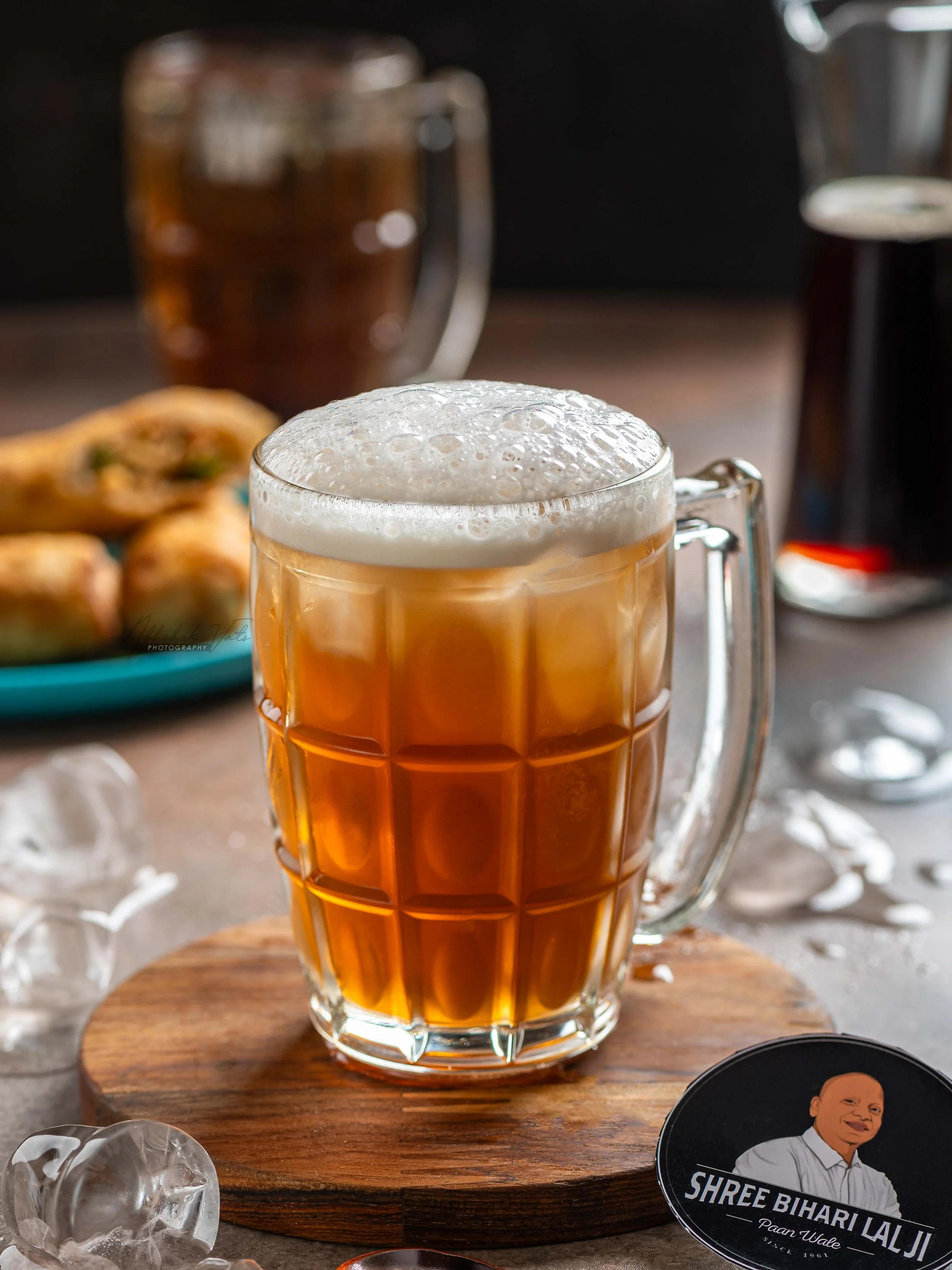 A glass mug filled with amber-colored beer topped with foam sits on a wooden board. In the background, there is a blurred dark object, possibly another drink. On the right, part of a round logo or sticker with a person’s image and text that reads "SH