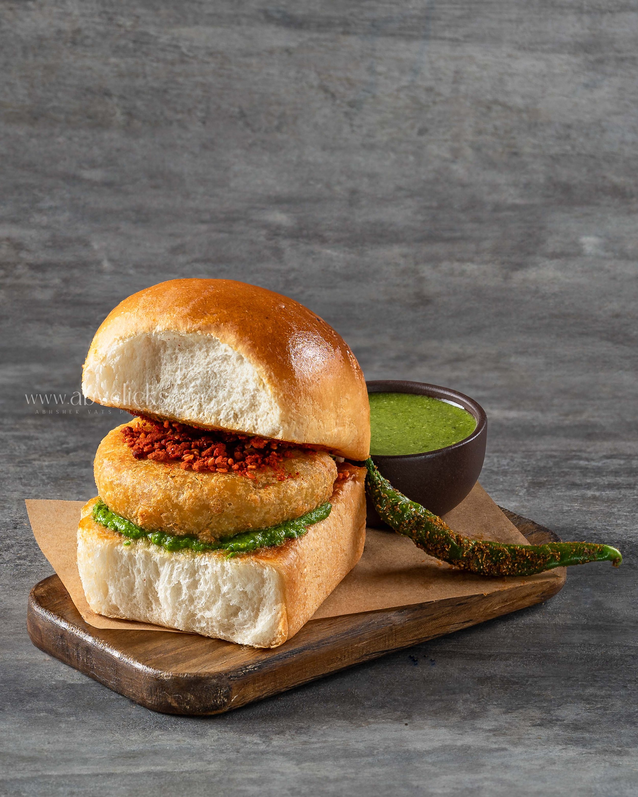 Close-up of a bun sandwich with a green sauce, placed on a wooden board with a bowl of green sauce and a grilled green chili on the side, against a gray textured background.