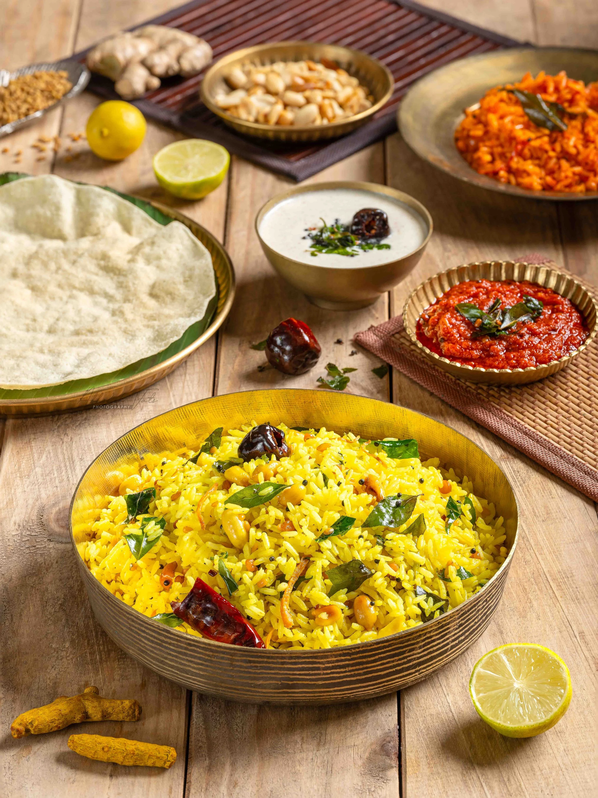 A variety of Indian dishes on a rustic wooden table, including a large bowl of yellow lemon rice with bay leaves and dried chilies, bowls of curry and chutney, naan bread, and garnished lemon halves.