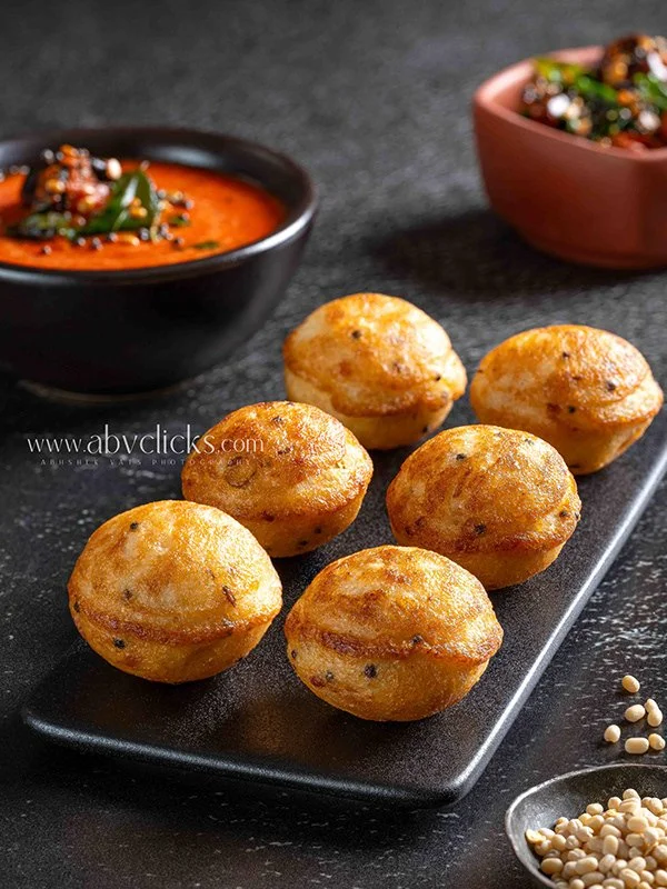 Six deep-fried spherical snacks on a black rectangular plate, with two bowls of dipping sauces in the background, one red and one black. The snacks appear golden-brown and crispy.