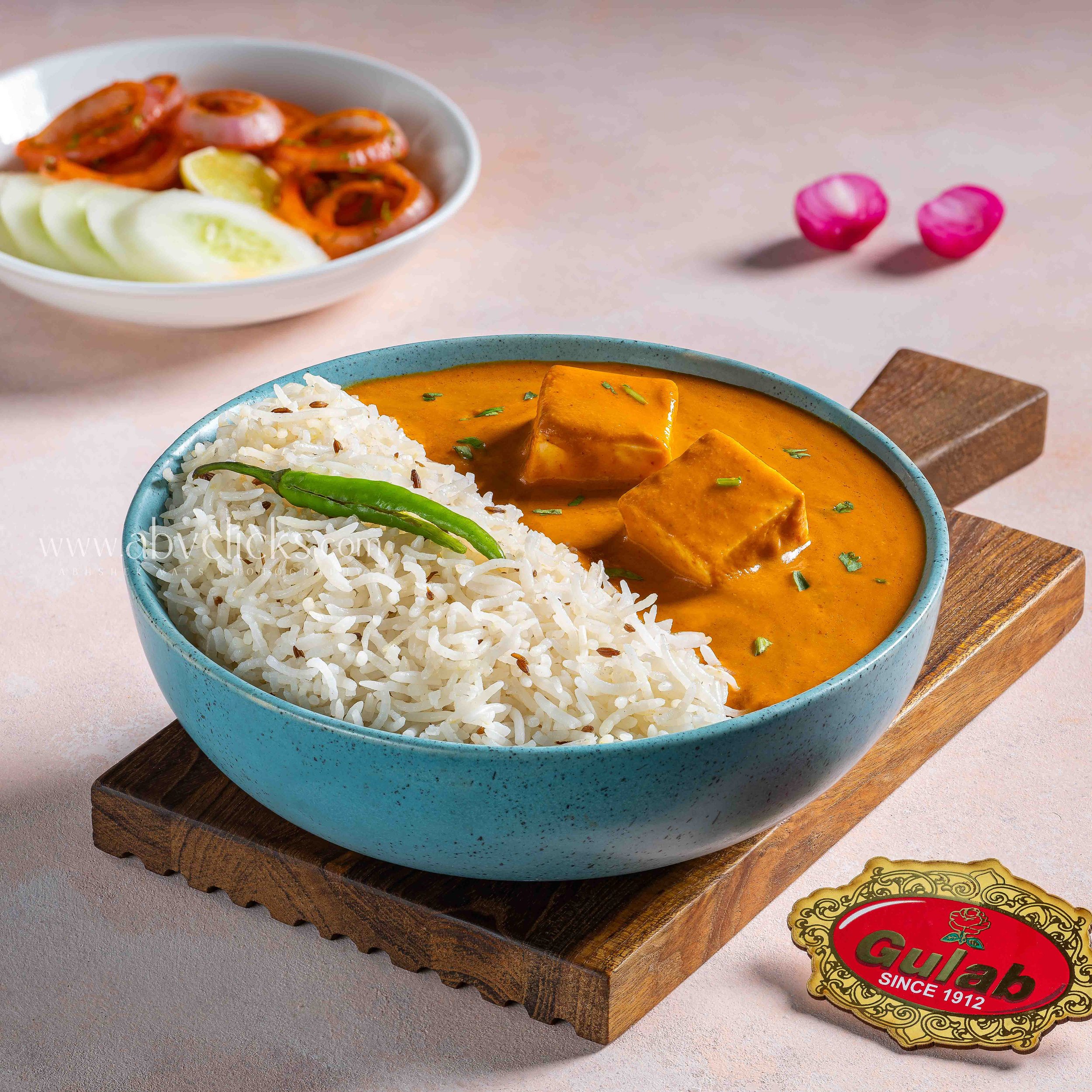 A bowl of Thai curry with rice and tofu, with a side of sliced cucumber, a lemon wedge, and two pink pickled onions, placed on a wooden serving board.