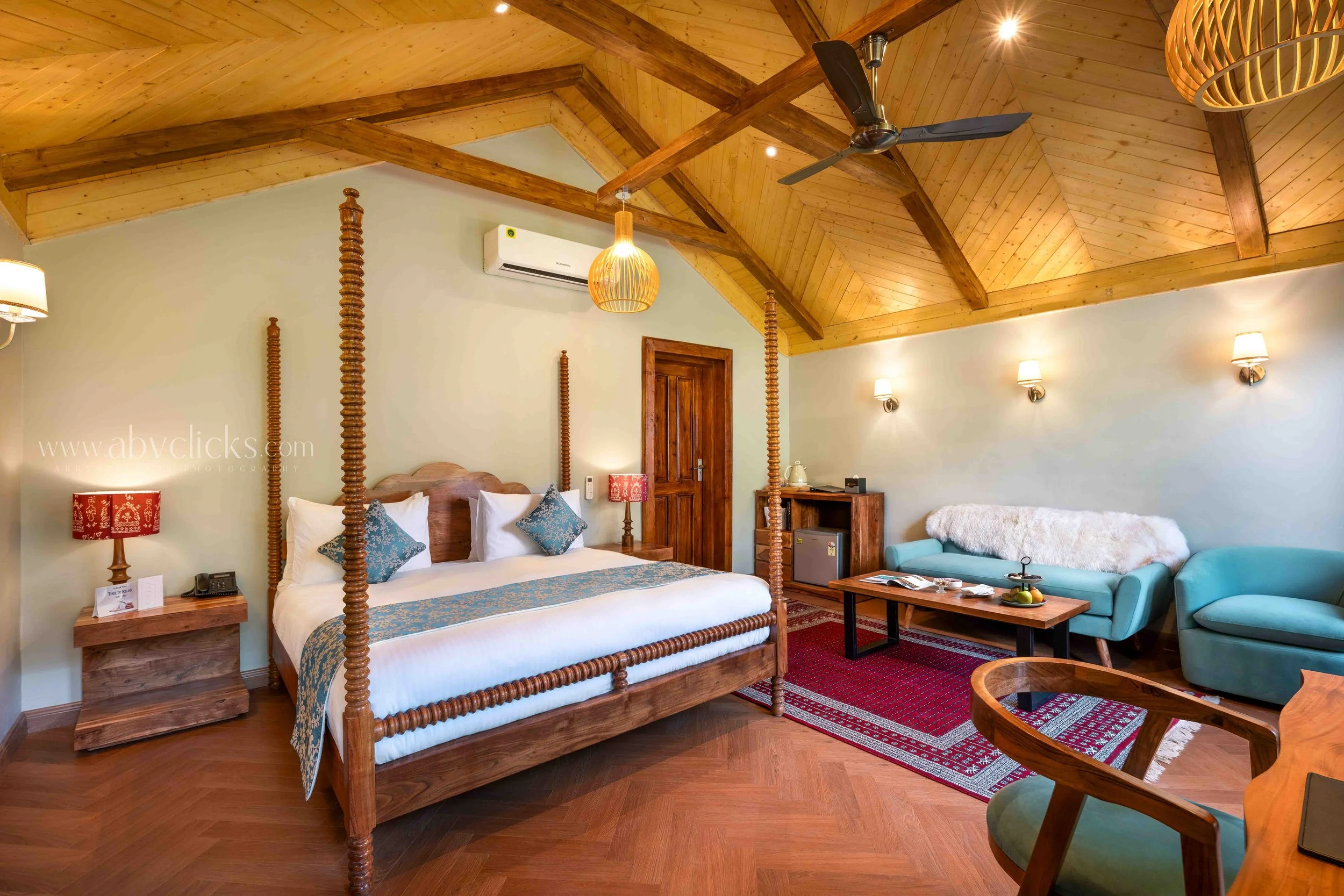A bedroom with a wooden four-poster bed, a turquoise sofa, and a ceiling fan, with wooden beams and warm lighting.