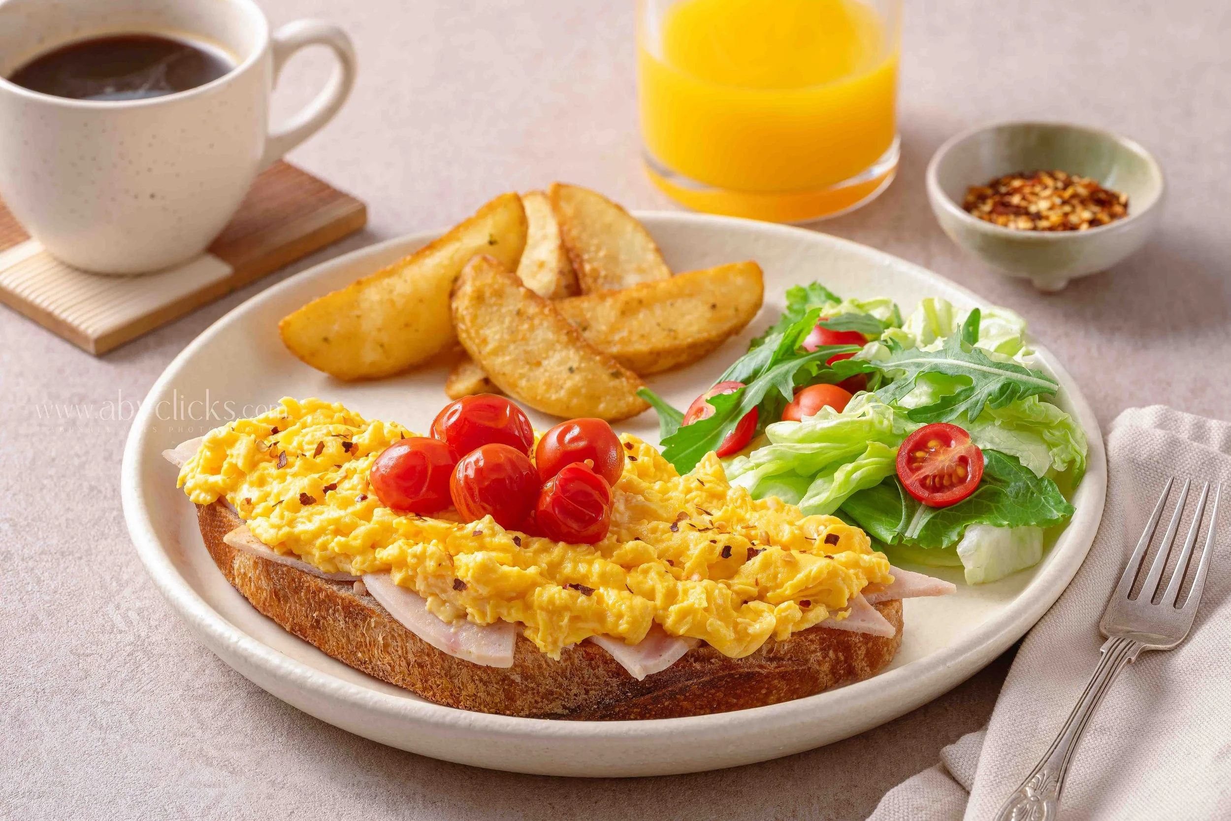 Plate with scrambled eggs, cherry tomatoes, lettuce, and toast with scrambled eggs and ham. Behind the plate, there is a cup of coffee, a glass of orange juice, and a small bowl of red pepper flakes.