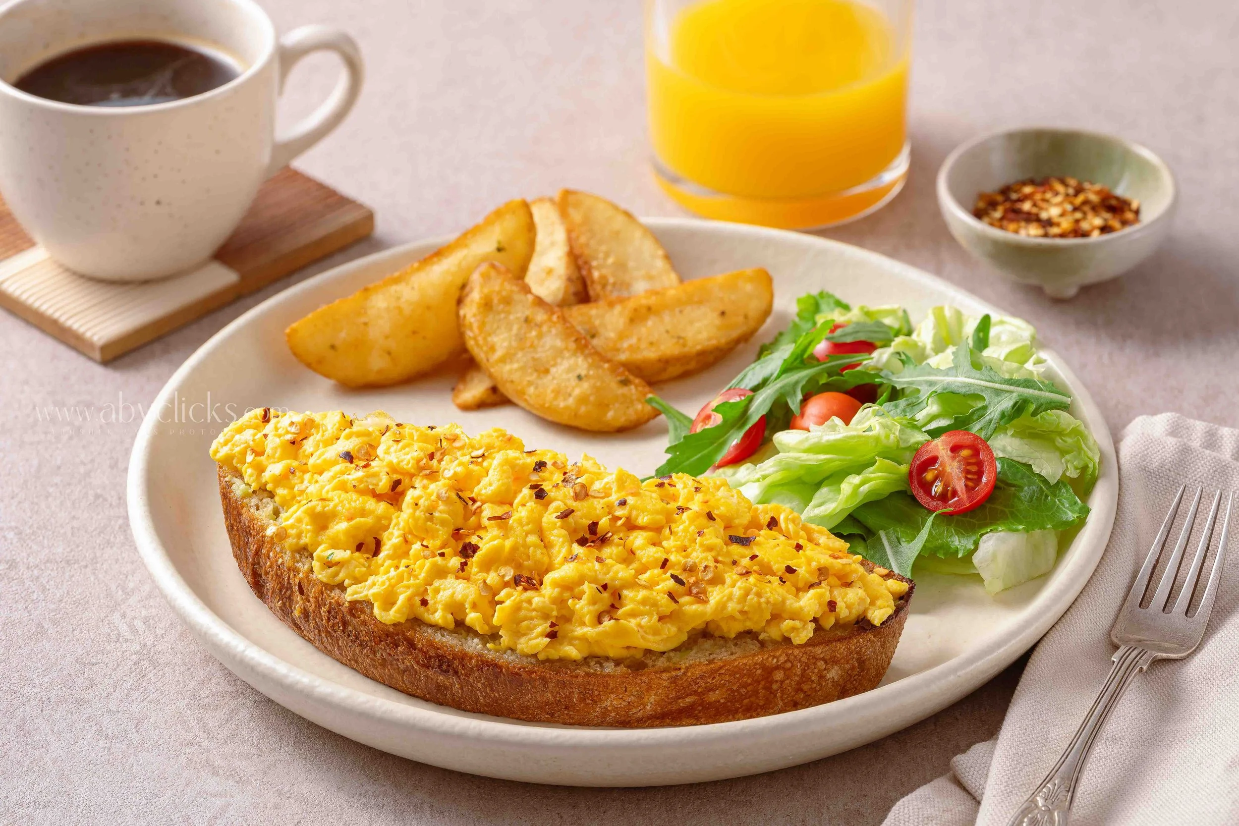 A breakfast plate with scrambled eggs on toasted bread, potato wedges, a side salad with cherry tomatoes and greens, a cup of coffee, a glass of orange juice, and a small dish of red pepper flakes.