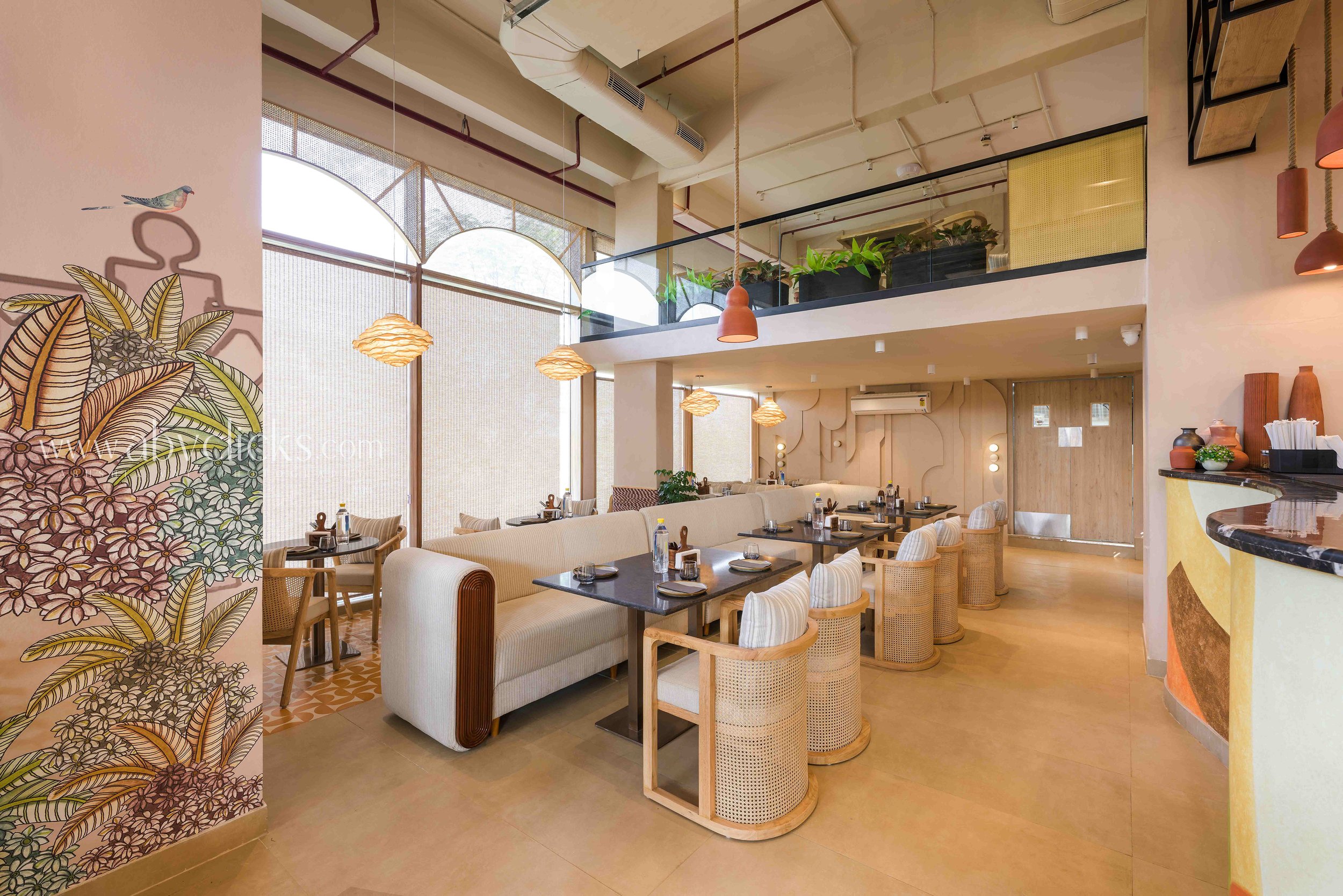 Interior of a modern restaurant with dining tables, chairs, large windows, hanging pendant lights, a decorative wall with floral mural, and indoor plants on an upper level.
