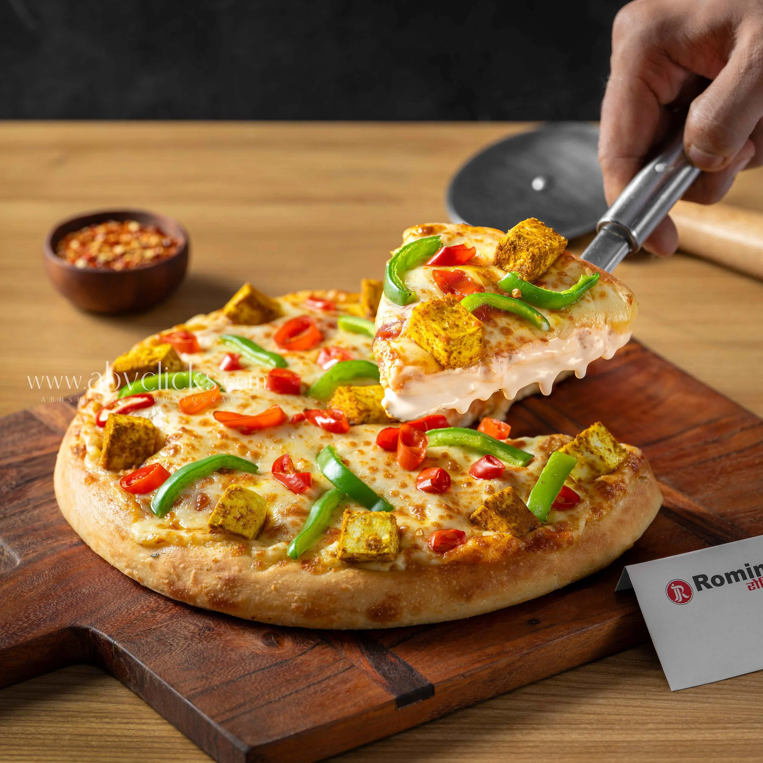 Close-up of a person using tongs to pick a slice of cheese pizza topped with green bell peppers and red chili peppers from a wooden pizza peel. In the background is a small bowl of red crushed pepper flakes on a wooden surface.