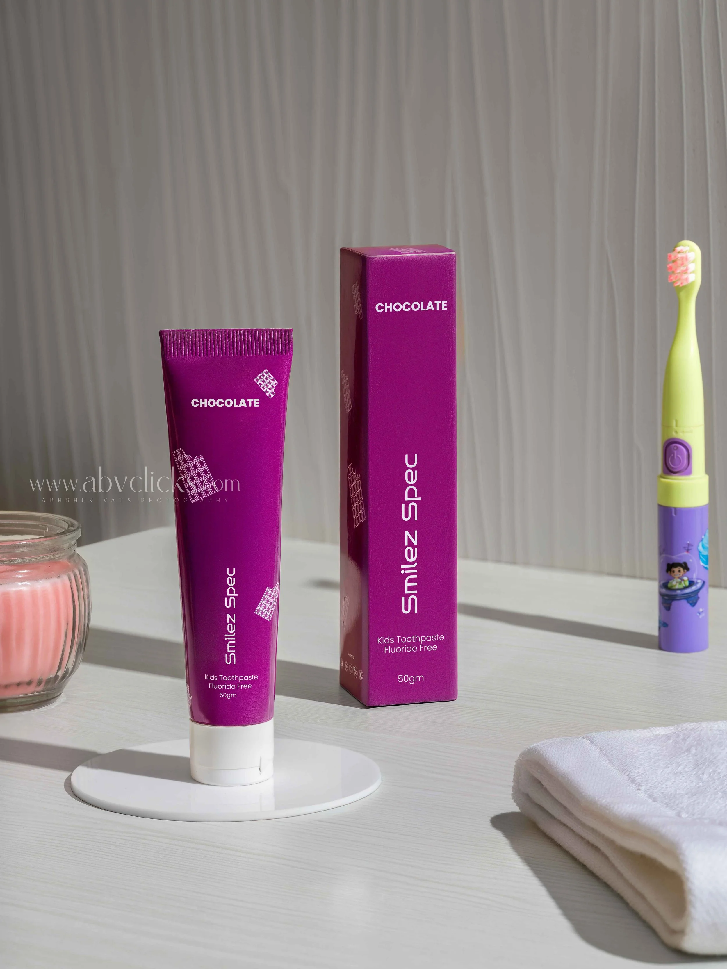 Pink kids toothpaste tube labeled 'Smilez Spelle Kids Toothpaste Fluoride Free 50gm' on a white display stand, with pink and purple packaging and a matching pink box behind it. To the right, a cartoon toothbrush with green and purple colors and a fol