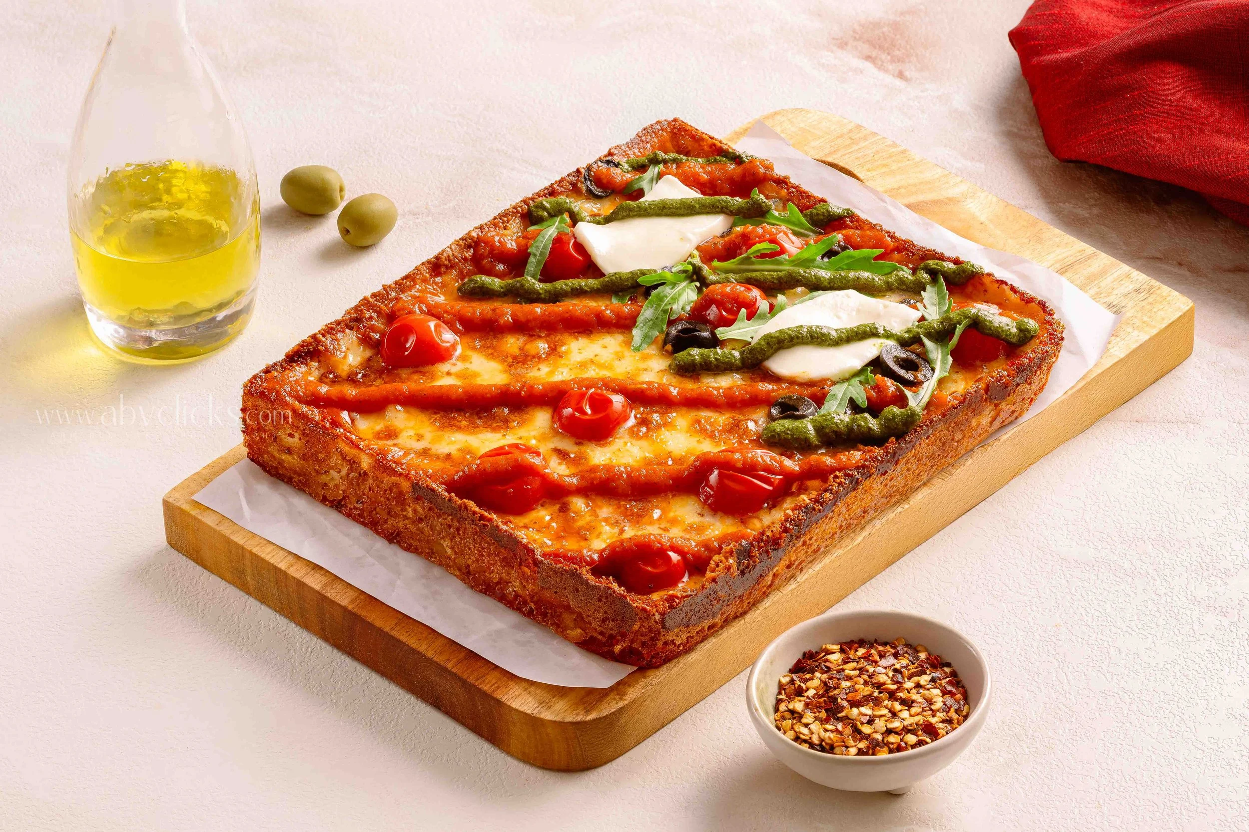 Italian-style rectangular pizza topped with cherry tomatoes, arugula, olives, green peppers, and cheese, arranged on a wooden serving board. A glass of olive oil, a small bowl of crushed red pepper flakes, and green olives are nearby.